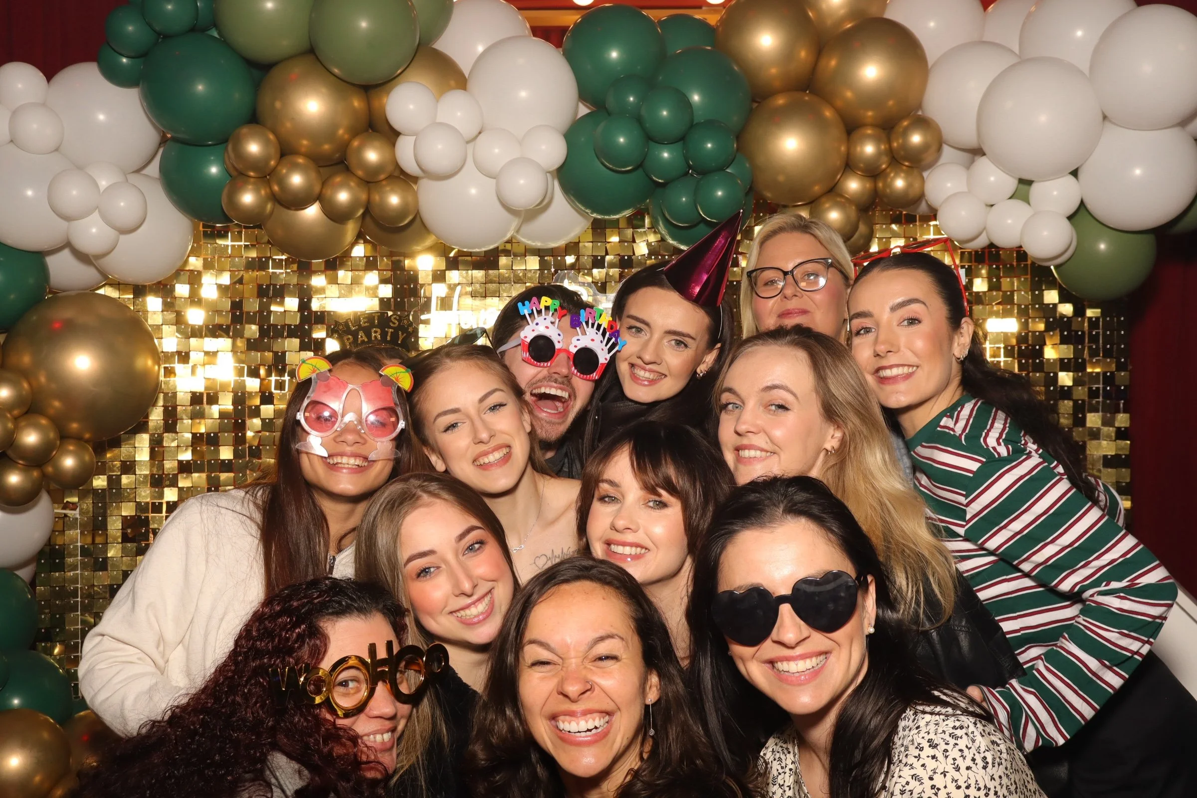 A group of diverse young adults celebrating at a party, smiling and wearing colorful accessories, with a backdrop of golden and white balloons and a sparkling gold wall.