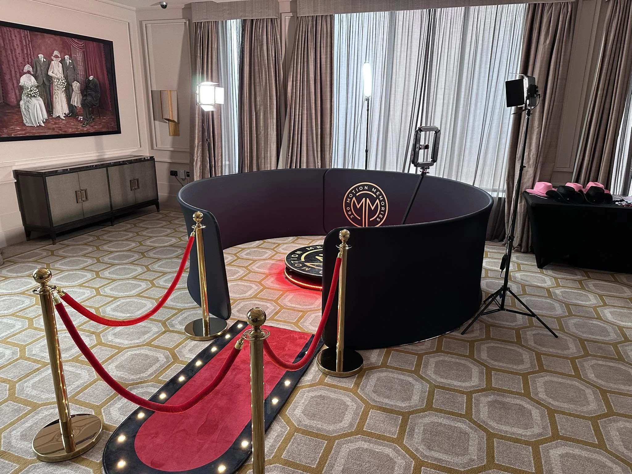 Indoor studio setup with a red velvet rope barrier, a circular black table with logo, studio lighting equipment, beige curtains, a framed painting of a vintage family portrait, and a cabinet on a patterned carpet.