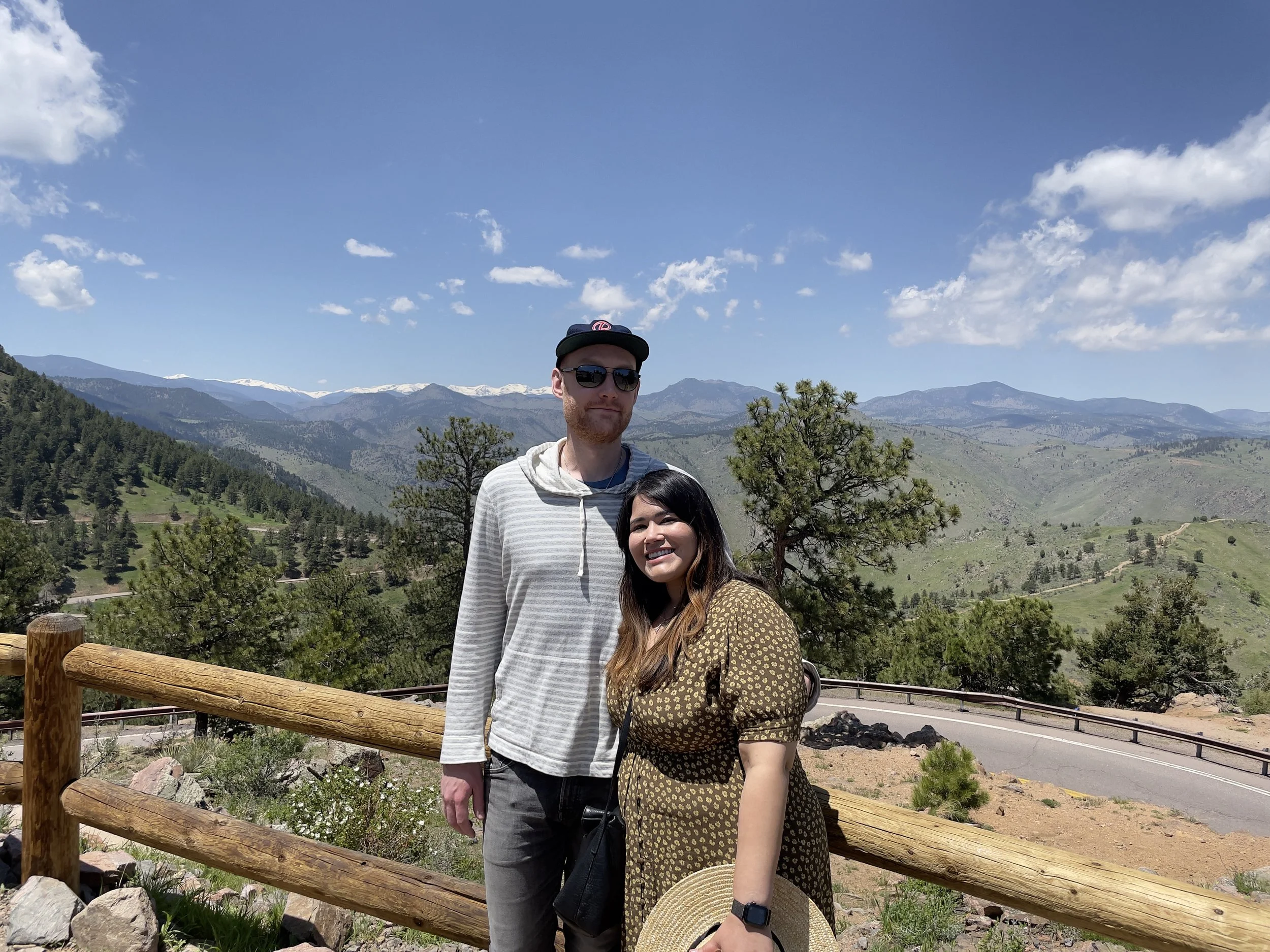 A man and woman stand together outdoors near a wooden fence, with a scenic mountainous landscape in the background. The man is wearing a light gray striped hoodie, sunglasses, and a cap, while the woman is dressed in a brown patterned dress and holding a wide-brimmed hat. They are smiling on a sunny day with a clear blue sky.