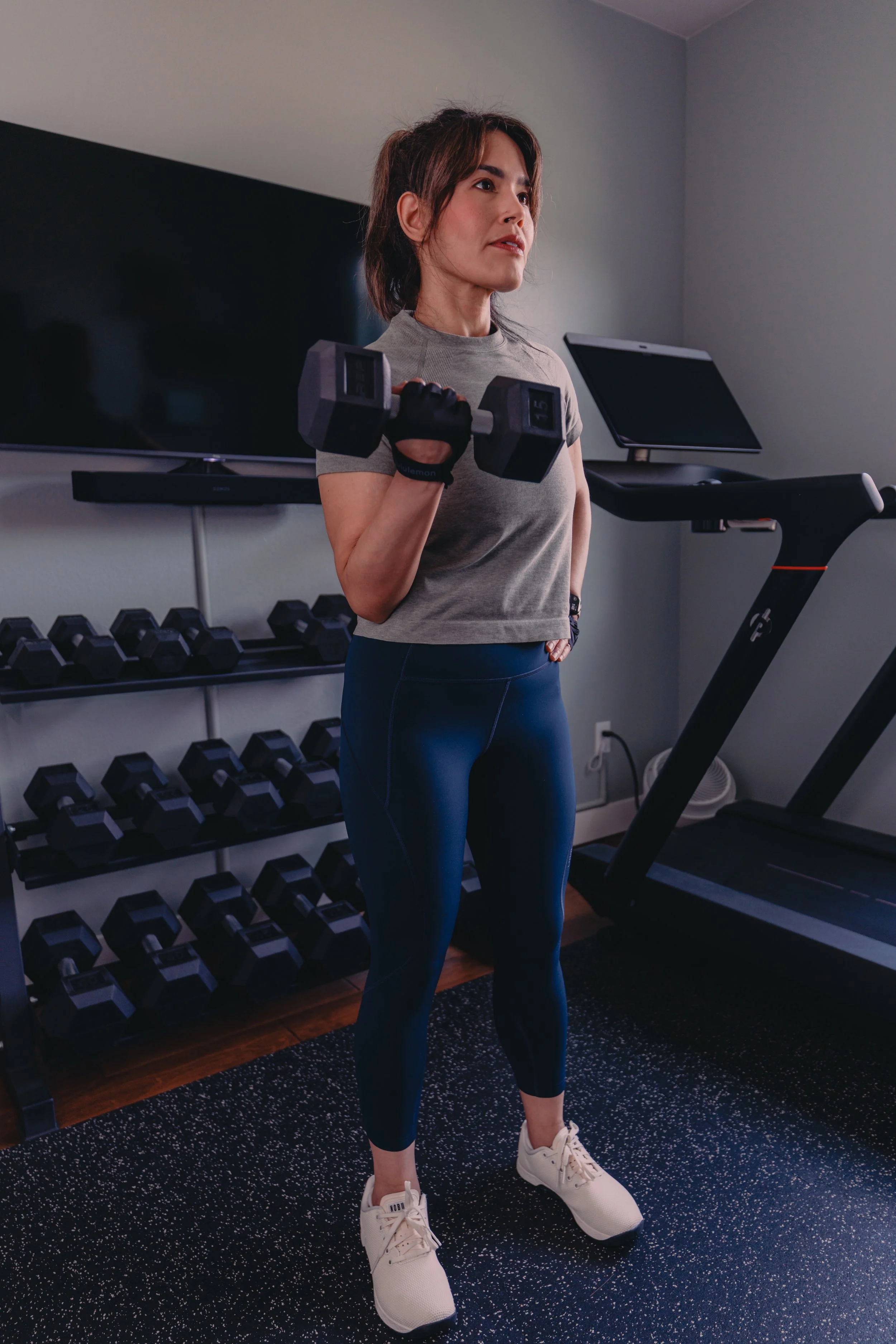 Woman lifting a dumbbell in a home gym, standing on a speckled black workout mat, with dumbbell weights on racks behind her, treadmill to her right, and a wall-mounted TV and computer screen in the background.
