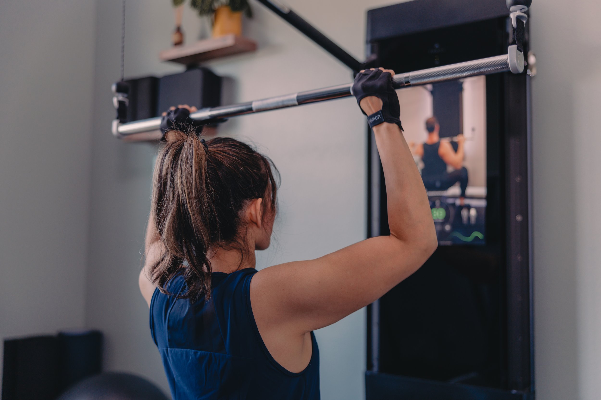 A woman with tied-back hair wearing a blue workout shirt lifting a barbell during a workout in a gym, with a mirror, black equipment, and a shelf with a plant in the background.
