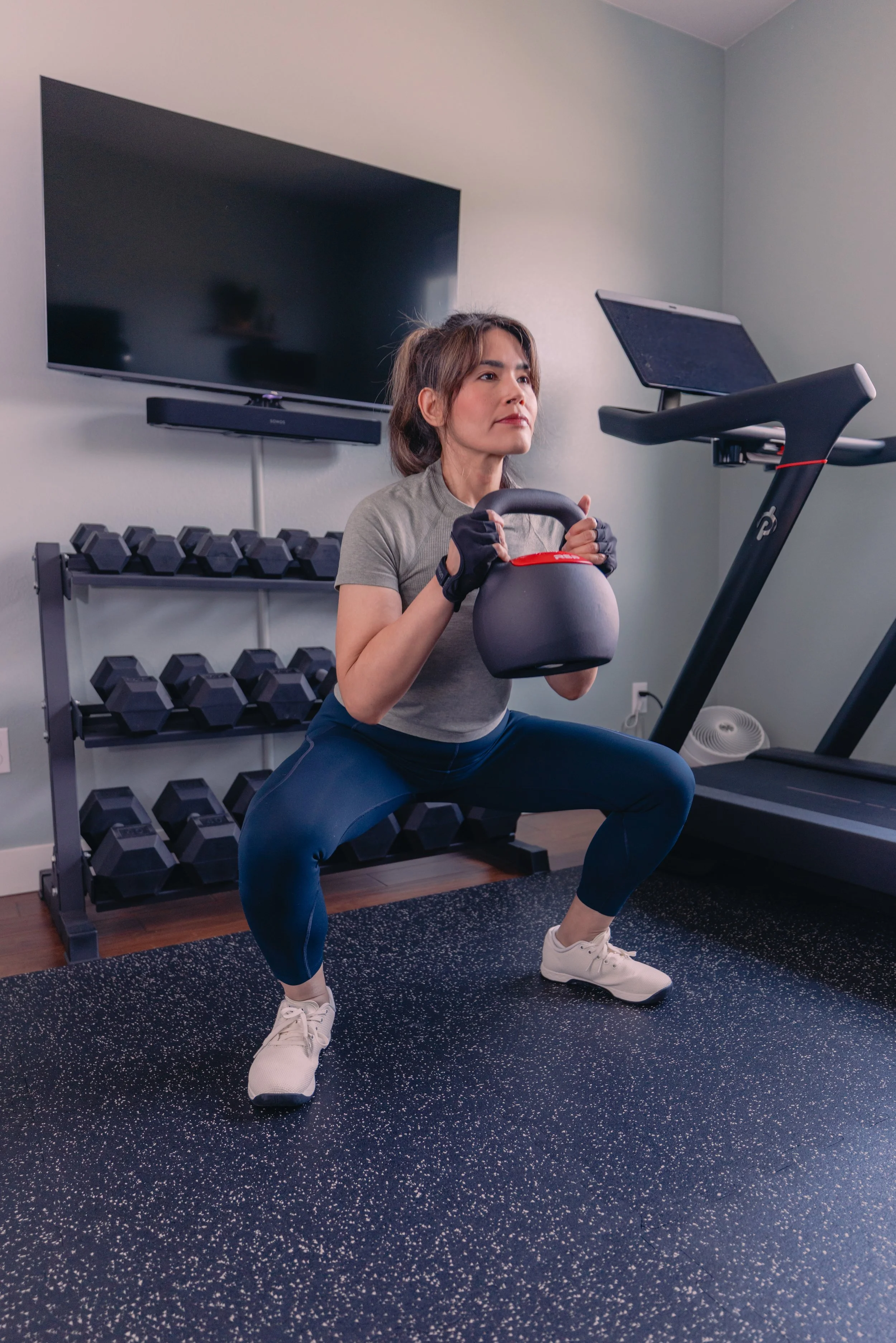 A woman in workout clothes, wearing gloves, squatting while holding a kettlebell, in a home gym with dumbbells on a rack and a treadmill nearby.
