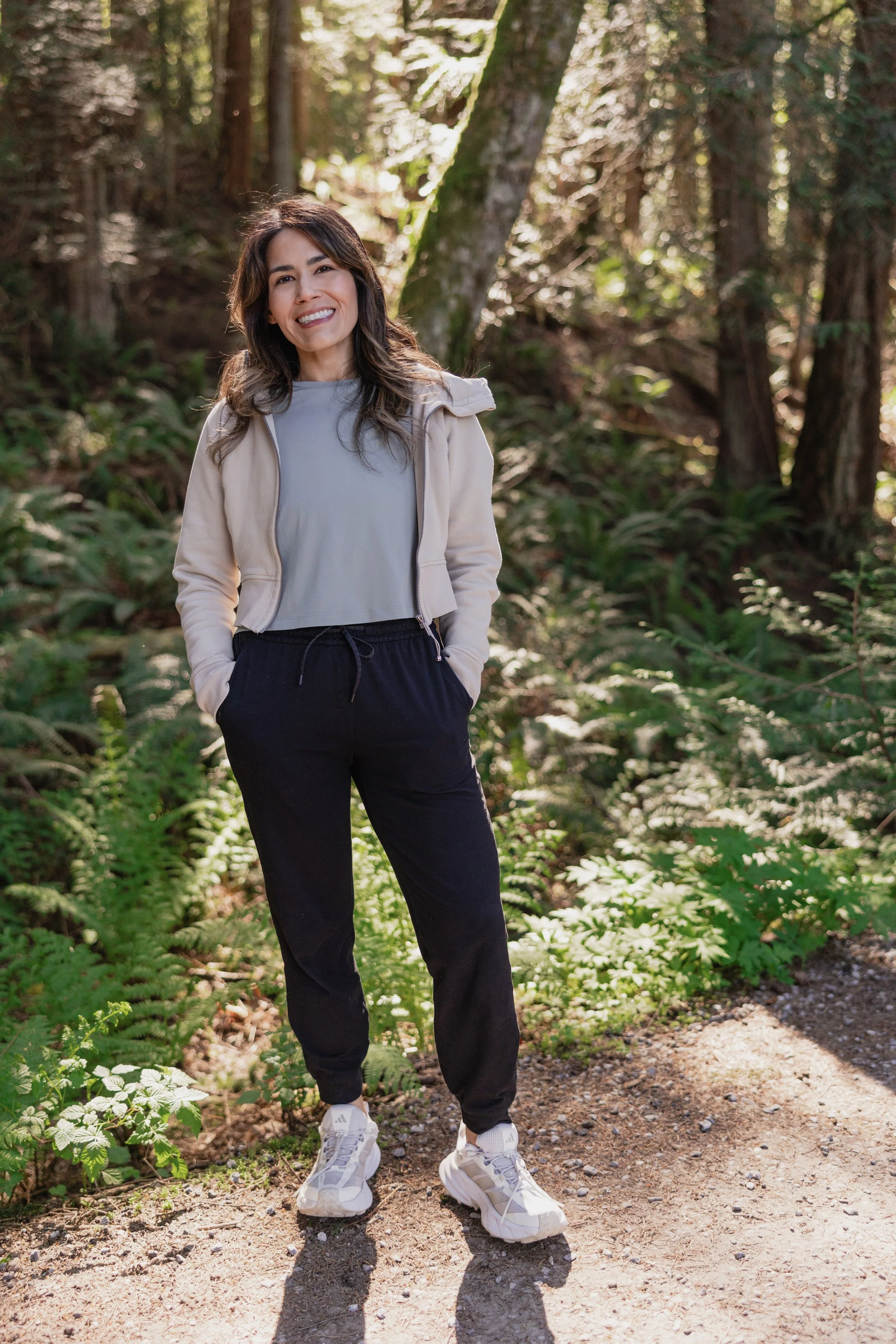 A woman standing on a dirt trail in a forest, smiling, wearing a light gray shirt, beige jacket, black pants, and white sneakers, with green foliage and sunlight filtering through trees in the background.