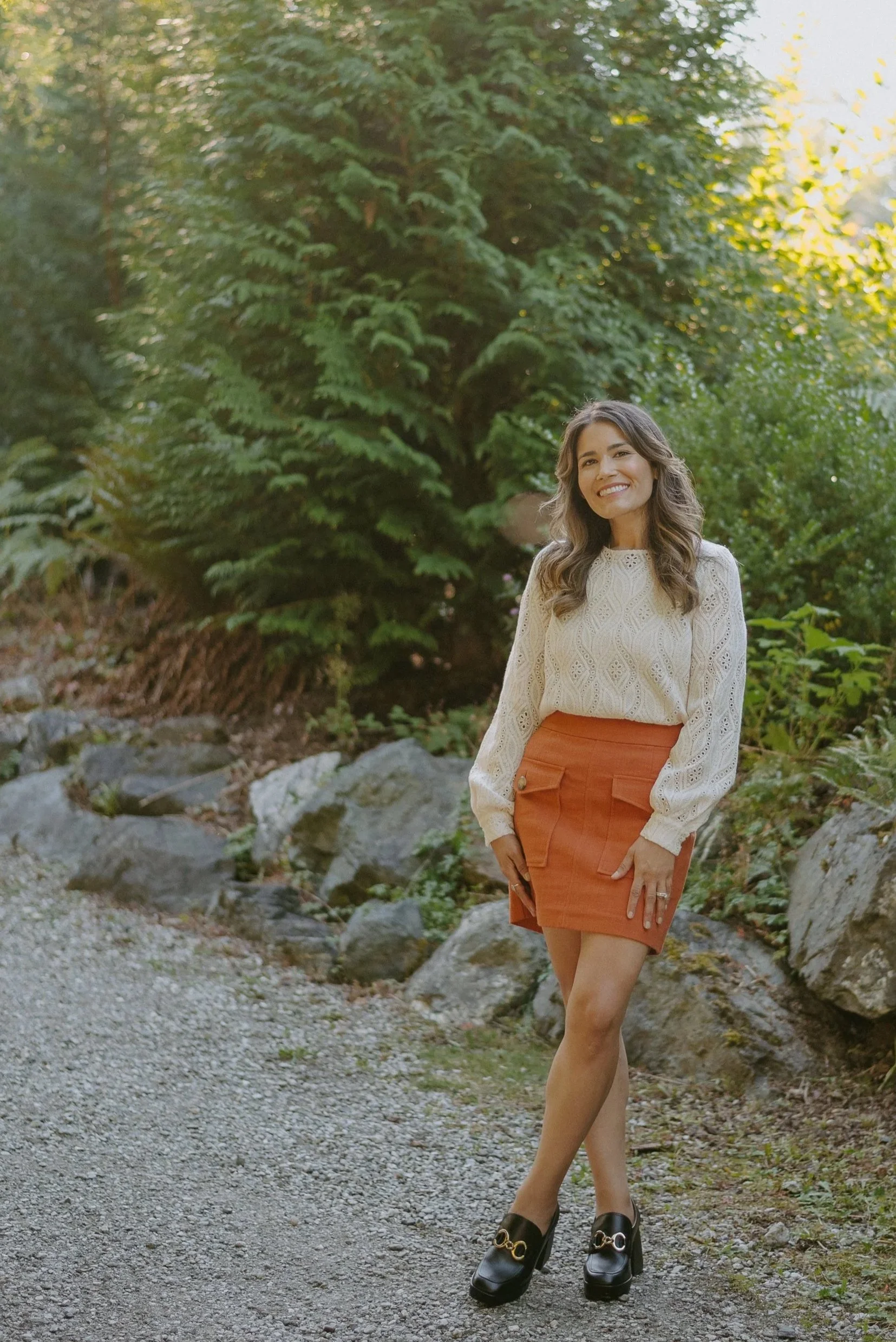A young woman with brownish hair, smiling, standing outdoors on a gravel path with rocks and lush greenery in the background, dressed in a cream eyelet sweater, orange skirt, and black loafers.