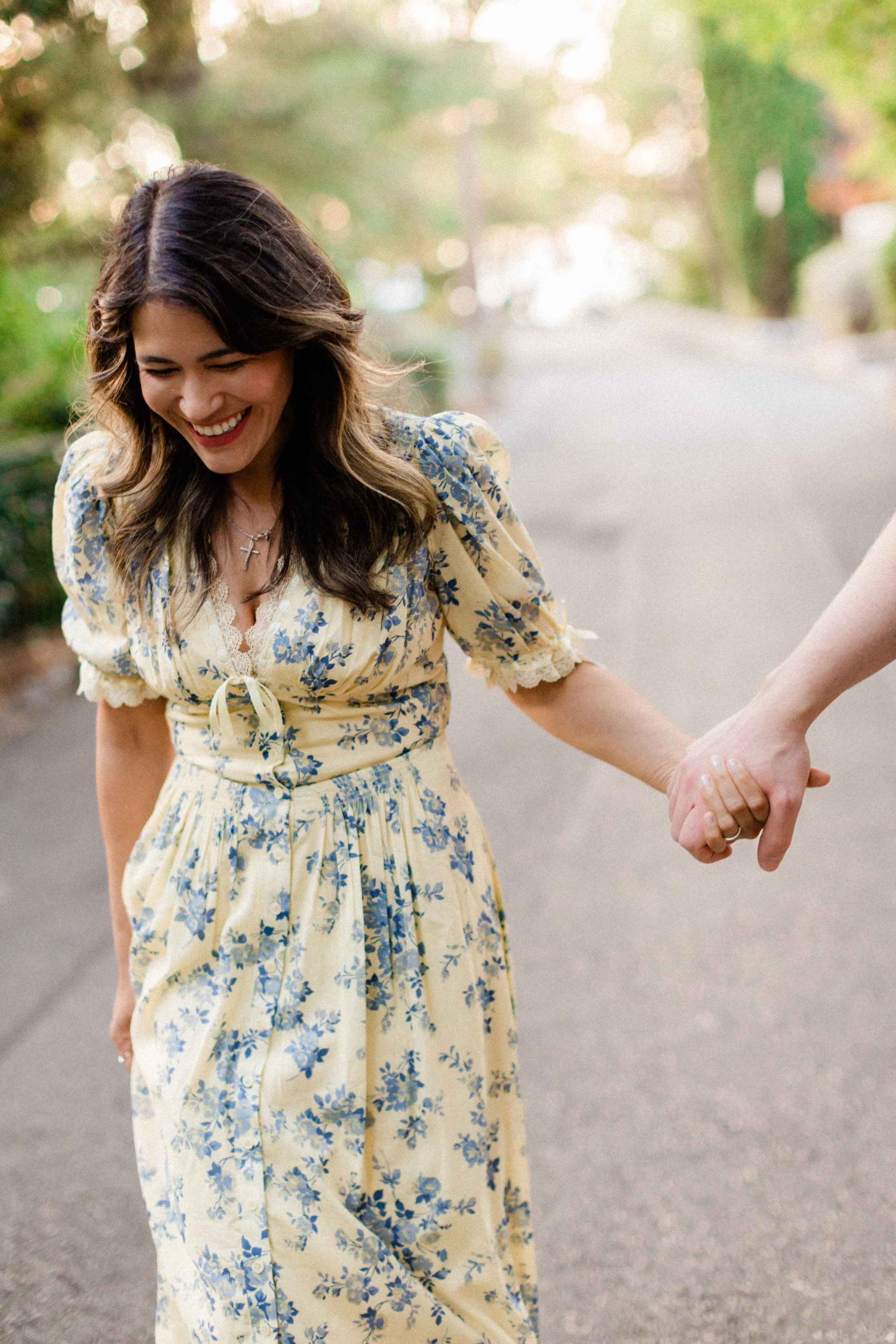 A woman in a yellow floral dress is holding hands with someone, smiling and looking down on a street with trees in the background.