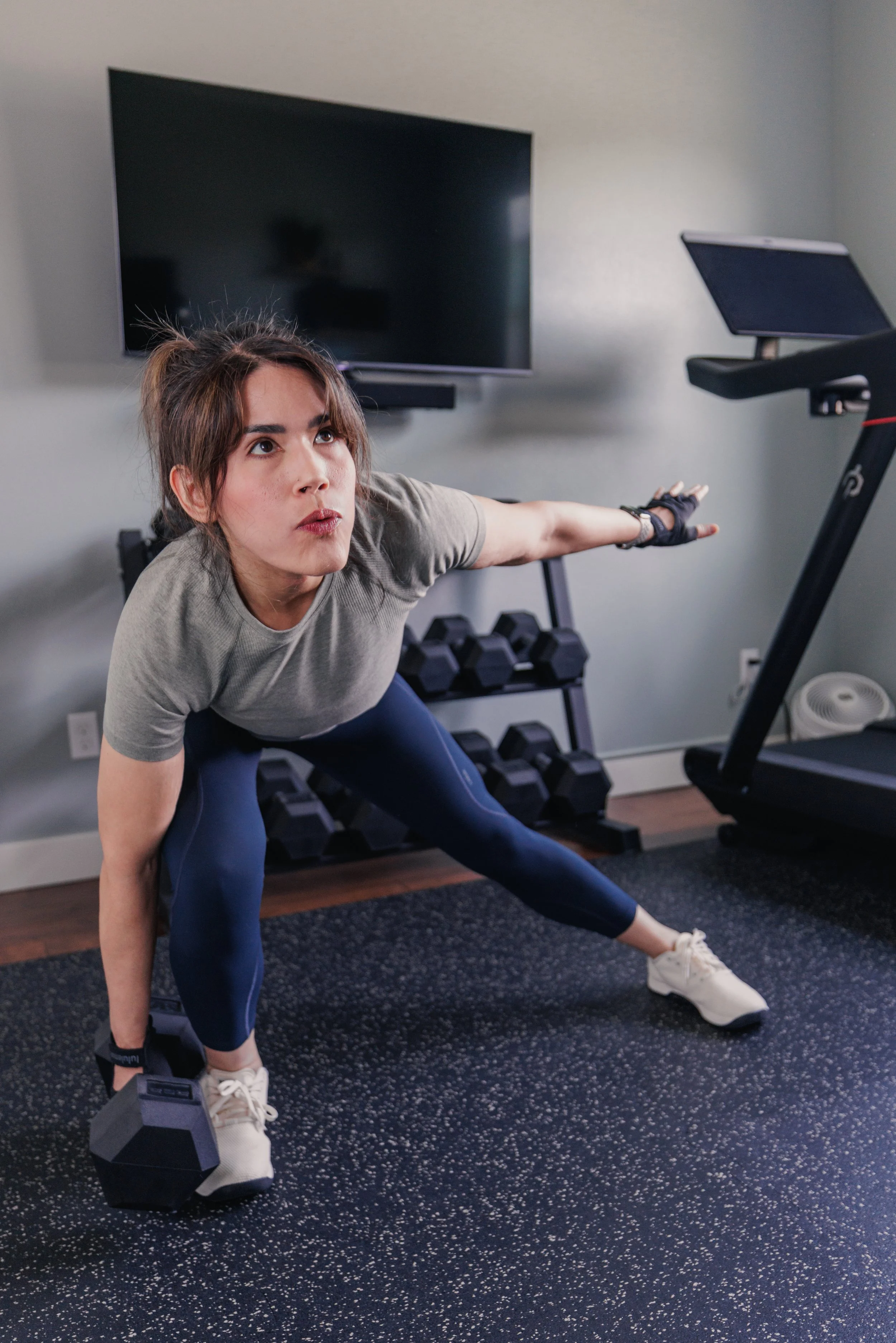 A woman doing a stretching exercise with dumbbells in a home gym, extending her right arm forward and her left hand on her left leg, surrounded by workout equipment and a large television on the wall.