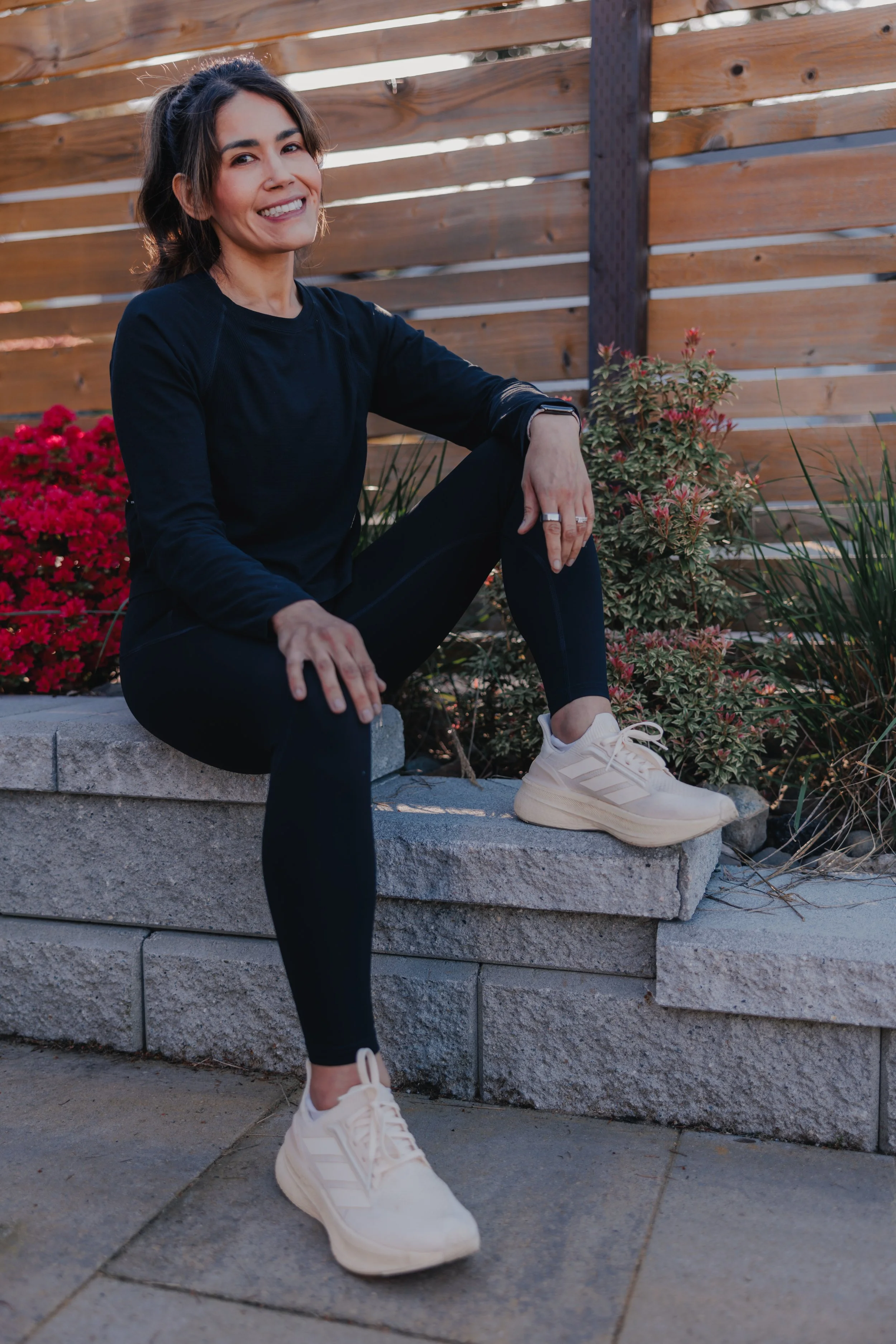 A woman with shoulder-length brown hair sitting on a stone ledge outdoors, smiling at the camera, wearing a black long-sleeve athletic shirt, black leggings, and cream-colored sneakers, with a wooden fence and pink flowers in the background.