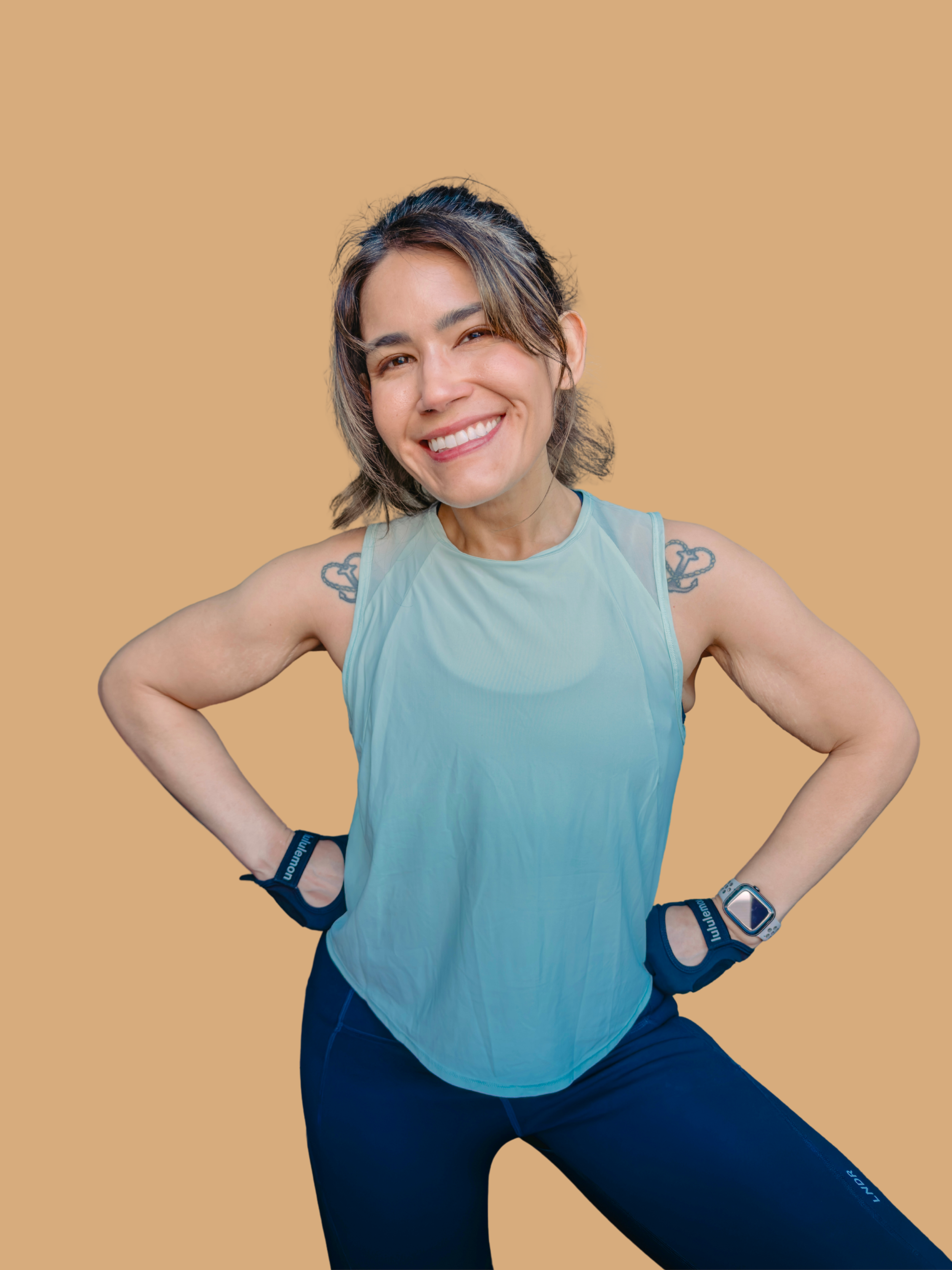 A woman smiling confidently with hands on hips, dressed in athletic wear including a light blue sleeveless top, dark blue leggings, fitness tracker, and wristbands, standing against a beige background.