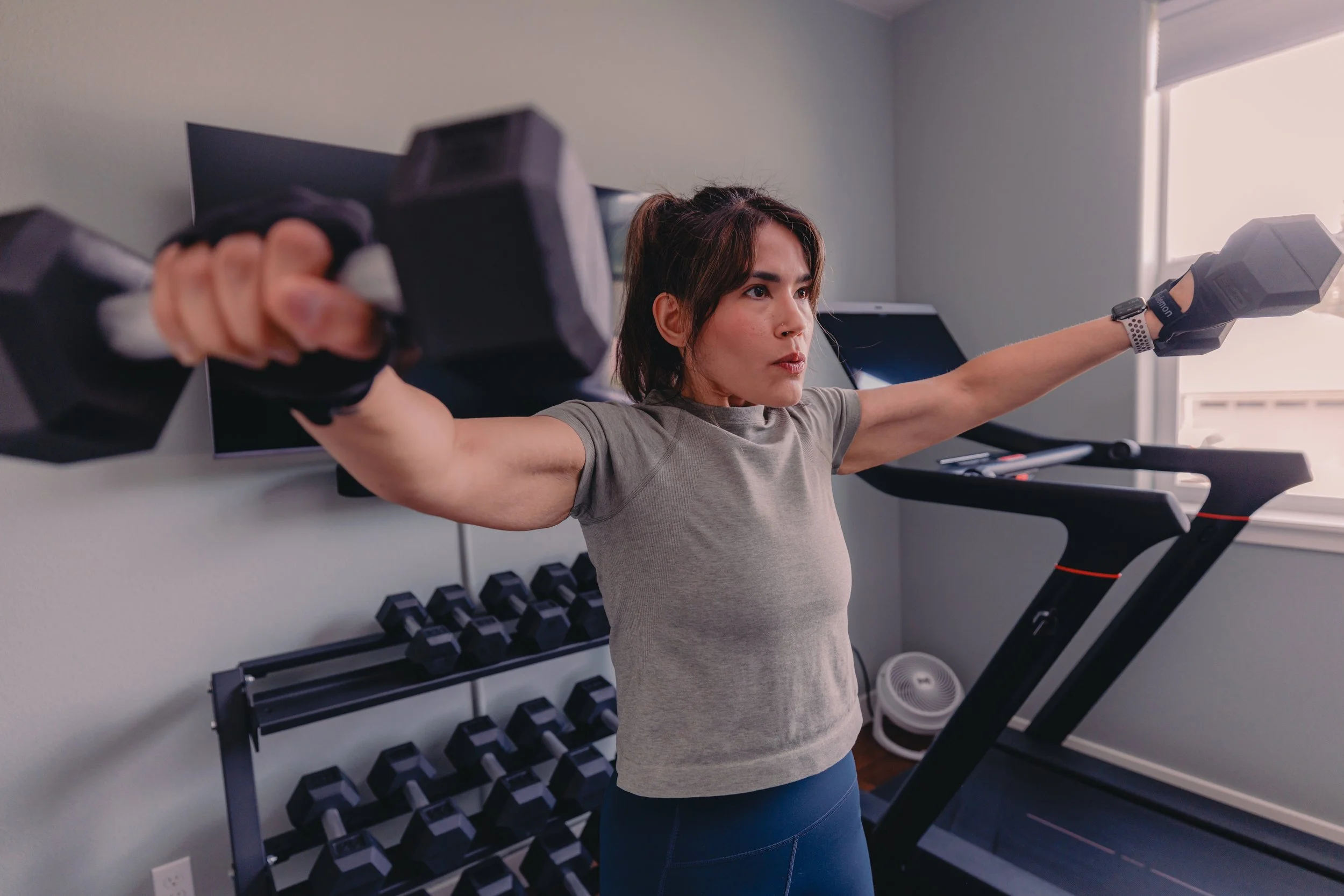 A woman in gray workout shirt and blue leggings is exercising on a treadmill in a home gym, holding dumbbells with arms outstretched. There are two black dumbbell racks behind her filled with black dumbbells, and a flat-screen TV on the wall. Natural light is coming through a window on the right.