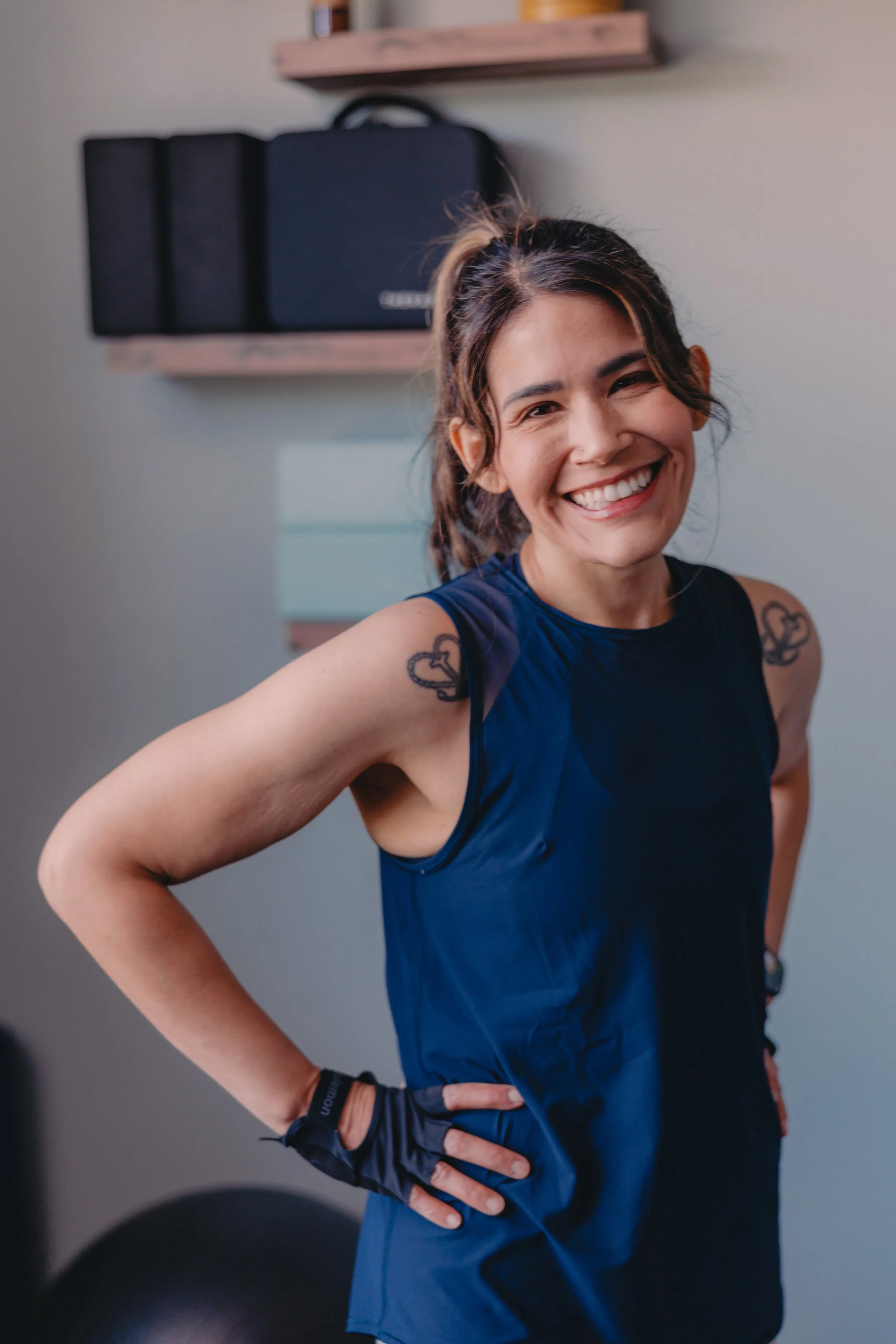 A woman smiling in athletic wear with tattoos on her arms, standing indoors in front of a wall with a wooden shelf and black equipment case.