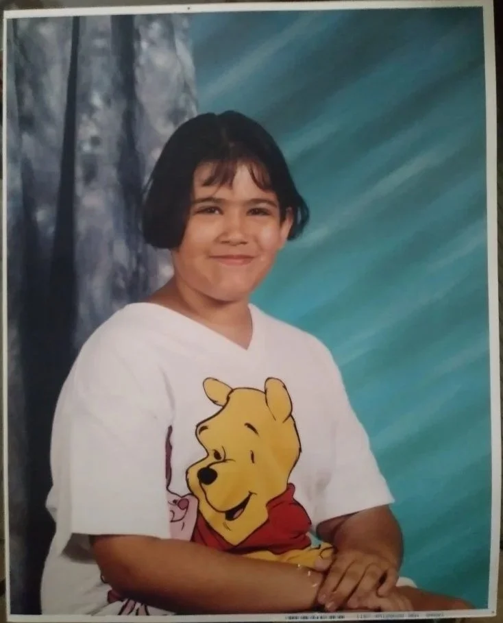 Child with short black hair smiling, wearing a white T-shirt with Winnie the Pooh graphic, sitting in front of a blue and gray backdrop.
