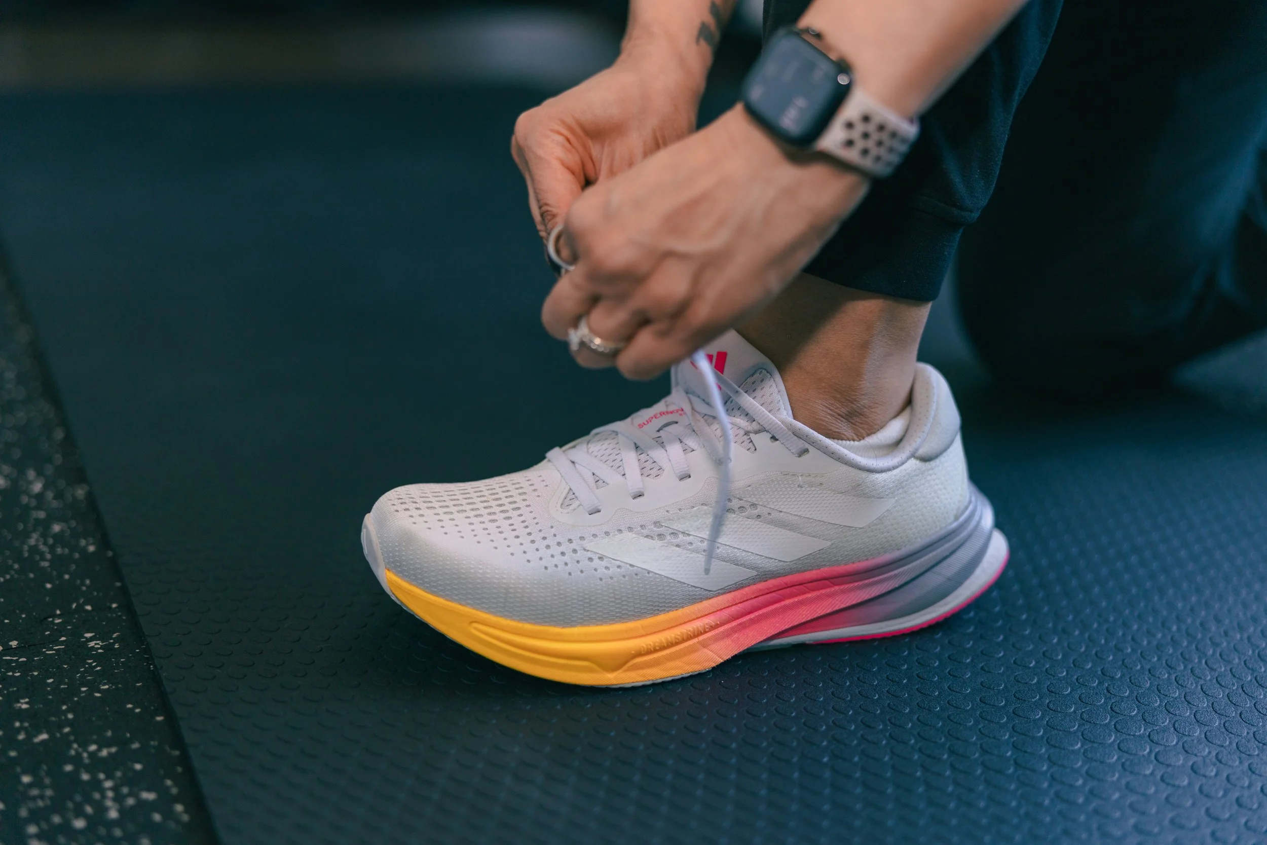 Person tying their shoe on a gym floor, wearing white athletic shoes with yellow and pink soles, black leggings, and a smartwatch on the wrist.