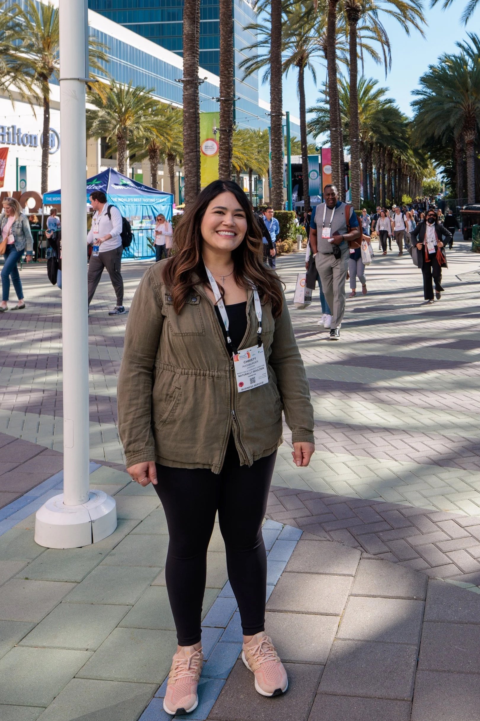 A woman with long dark hair smiling at the camera, standing outside a convention center with palm trees and people walking by.