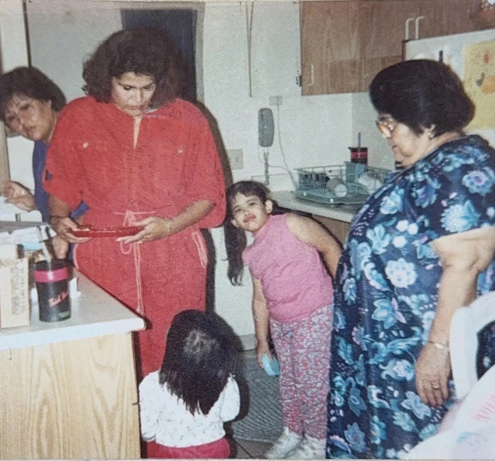 Four women and two children in a kitchen. One woman in a red dress is holding a plate, another woman with glasses and a blue floral dress is looking down, and the third woman with a pink dress is smiling and leaning forward. The children are seated at a table with one girl looking up at the women and the other girl bending forward. The kitchen has cabinets, a dish rack, and a microwave visible.