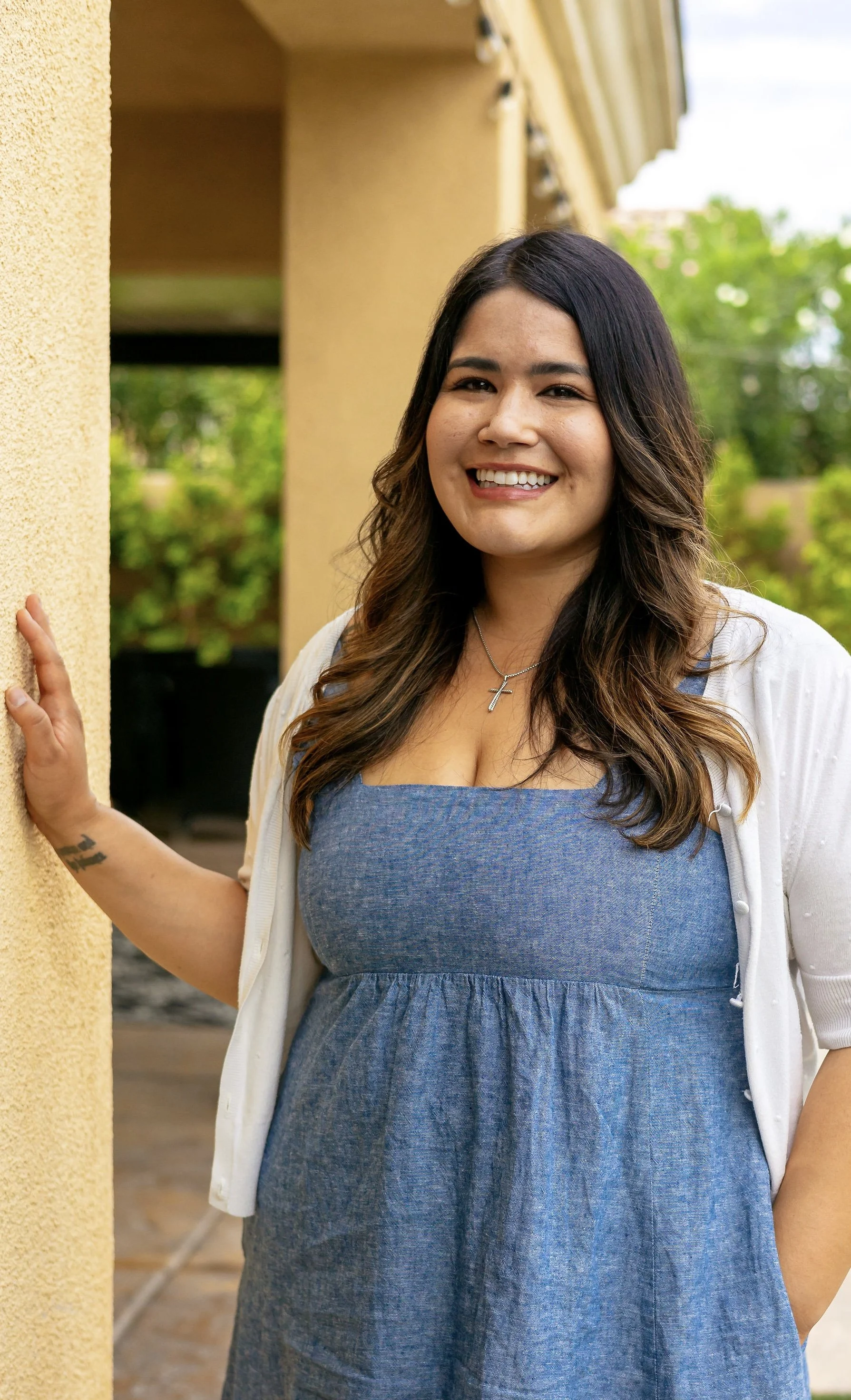 A woman with dark hair and a cross necklace standing outdoors, smiling, and touching a yellow wall.