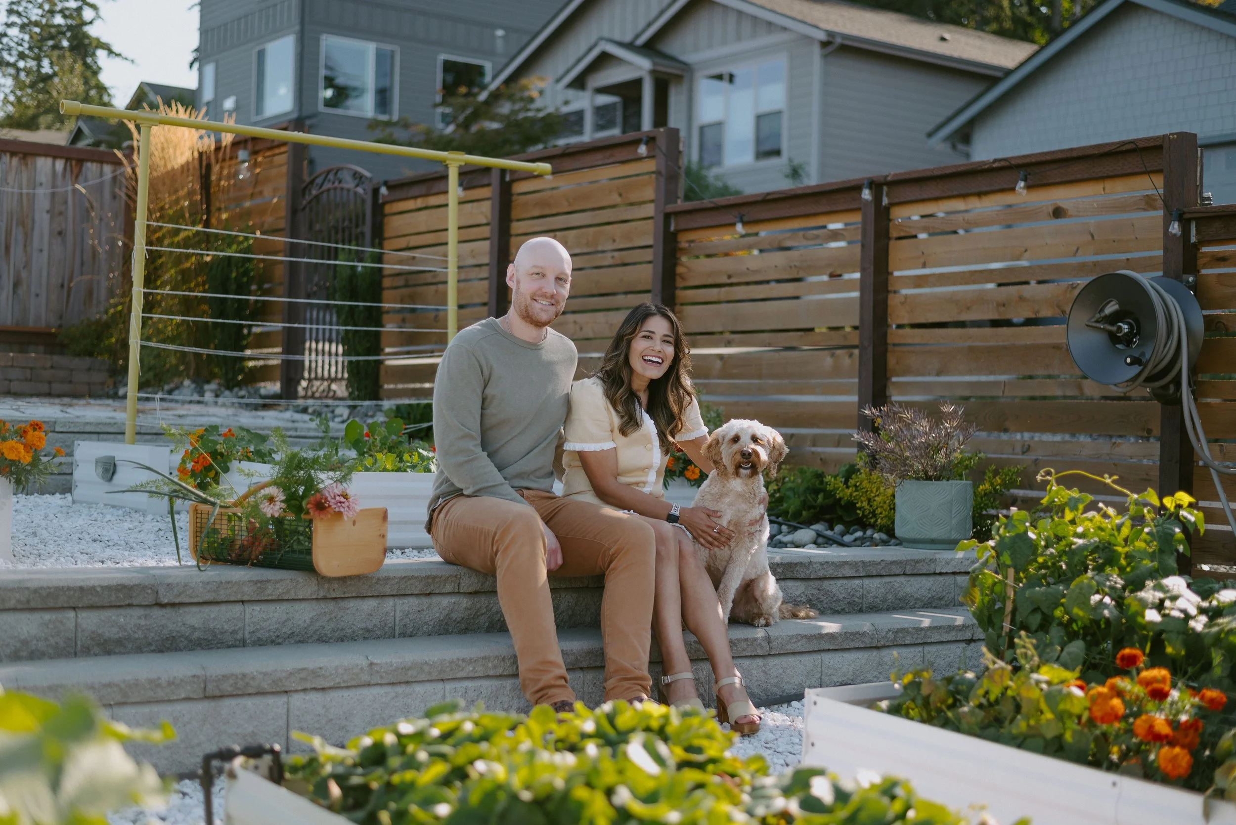 A happy couple with a dog sitting on outdoor steps in a backyard garden, smiling at the camera.