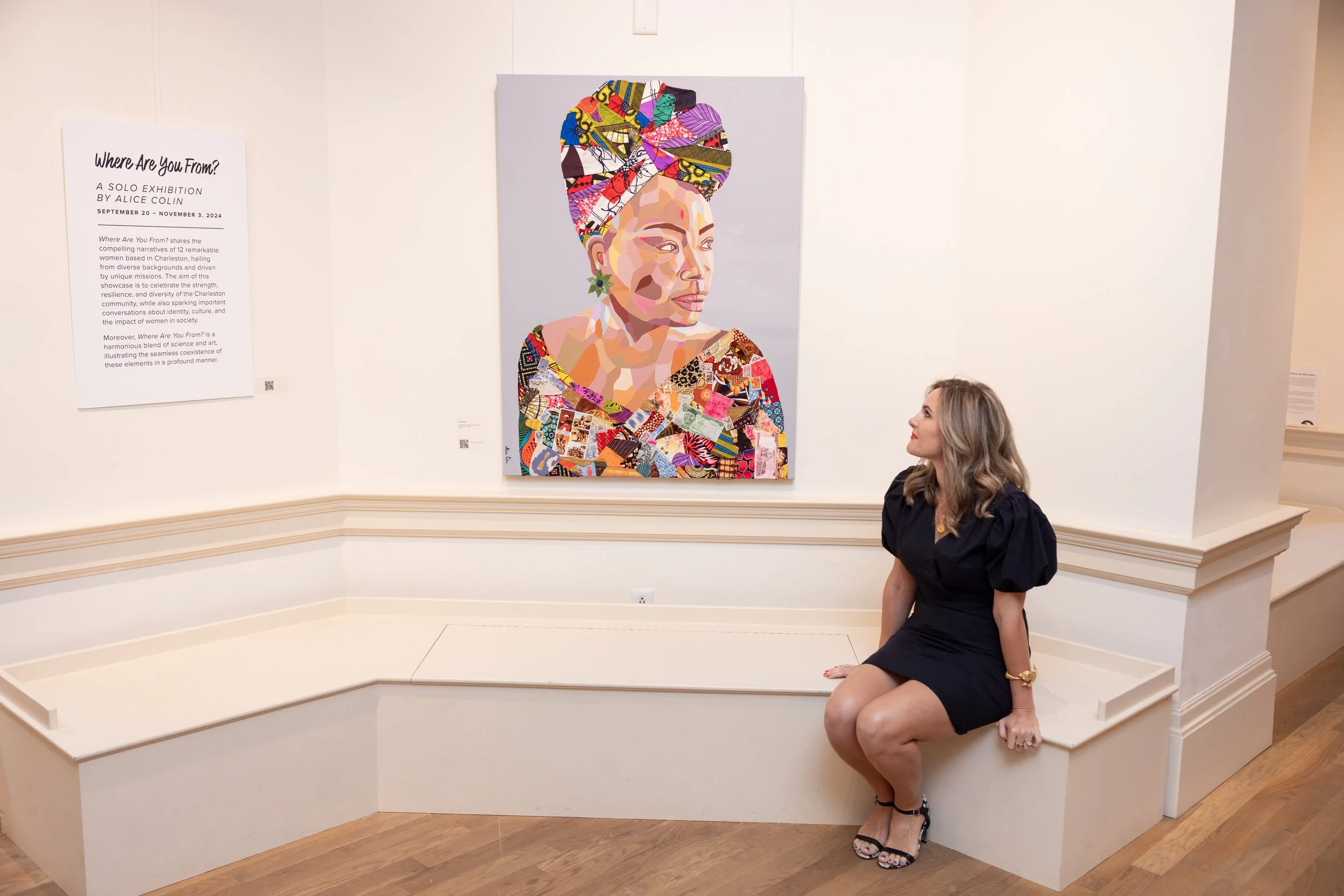 A woman with long, wavy gray hair sitting on a white bench in front of a colorful portrait of a woman with an elaborate, patterned headwrap. The woman in the foreground is wearing a black dress and sandals, and is looking at the portrait.