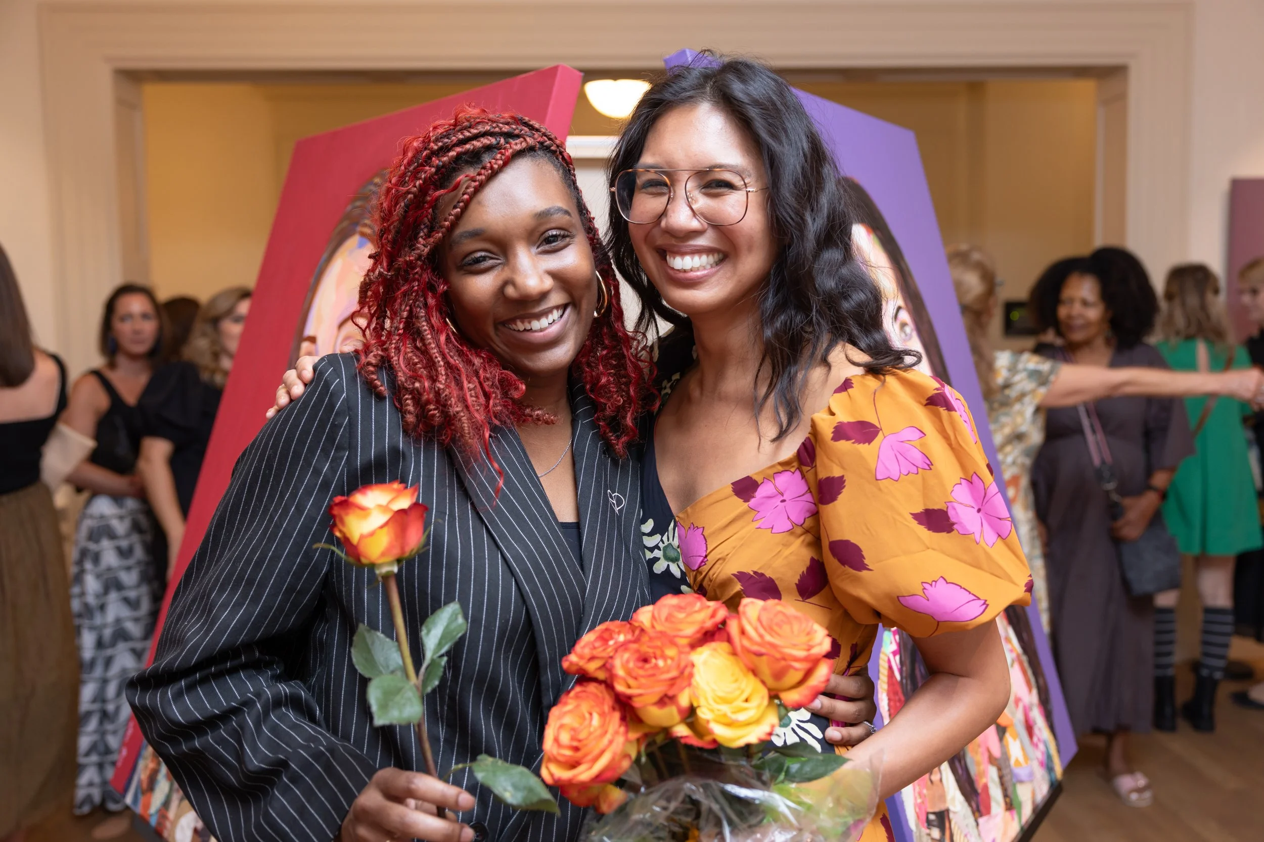 Two women smiling, holding colorful roses, at an indoor event with other people in the background.