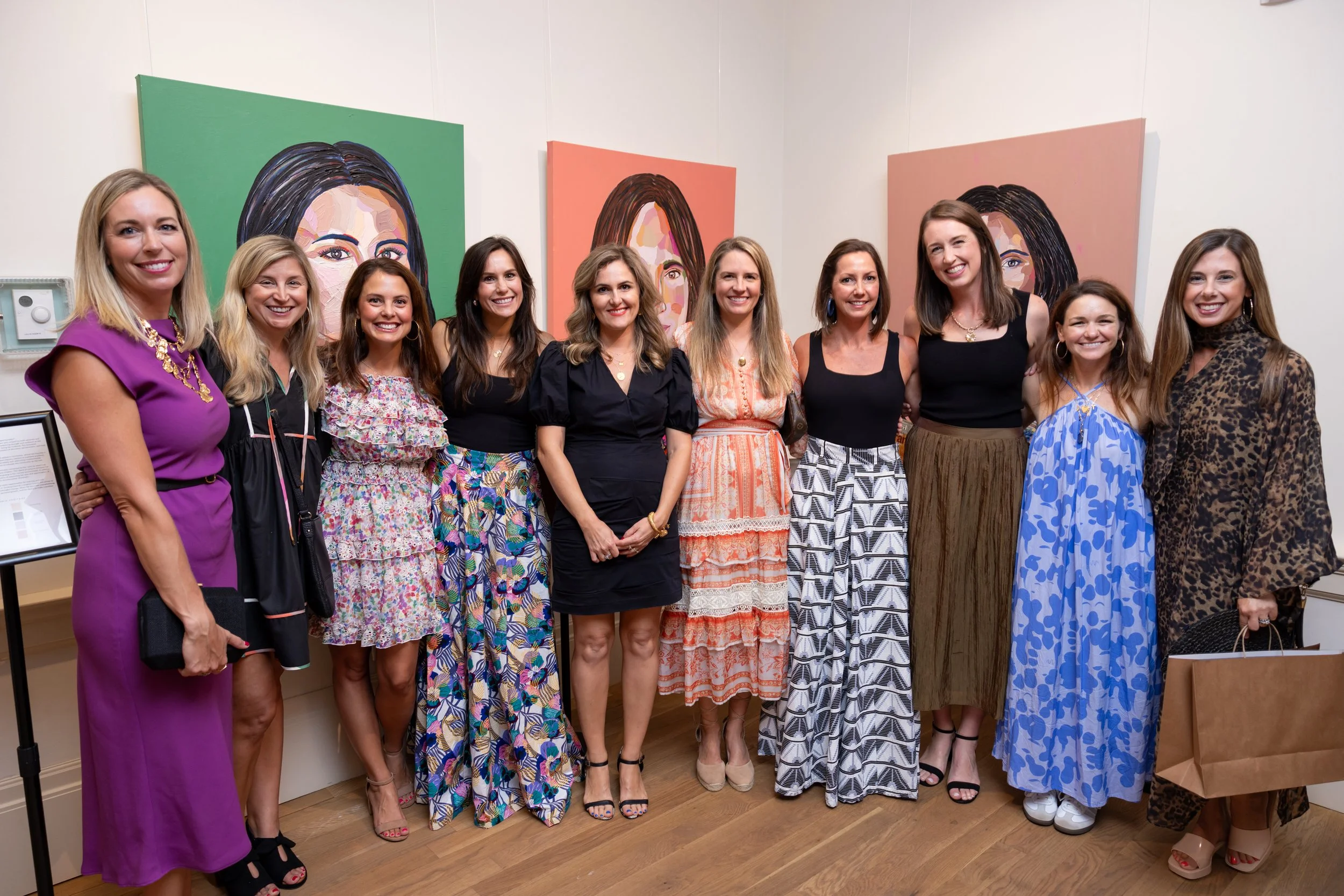 A group of twelve women standing together indoors at an art gallery, smiling for a photo in front of colorful portraits of women painted on the wall behind them.