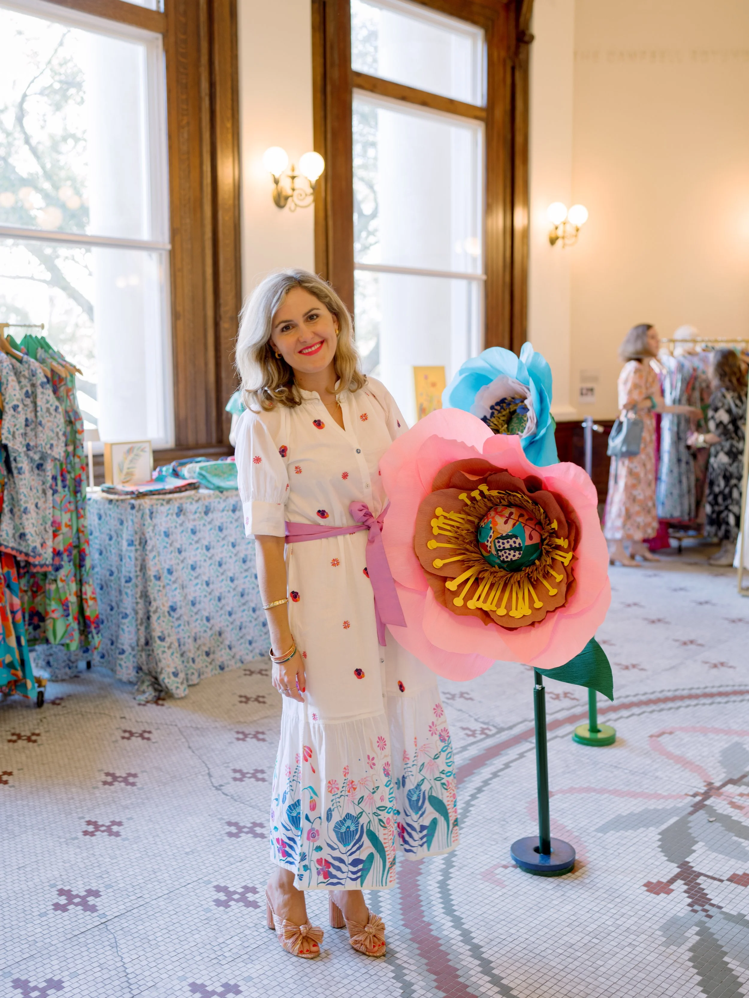 A woman in a white dress with colorful floral patterns, tan shoes with bows, stands next to a colorful paper flower sculpture in a room with large windows, wooden trim, and other women browsing clothing racks in the background.