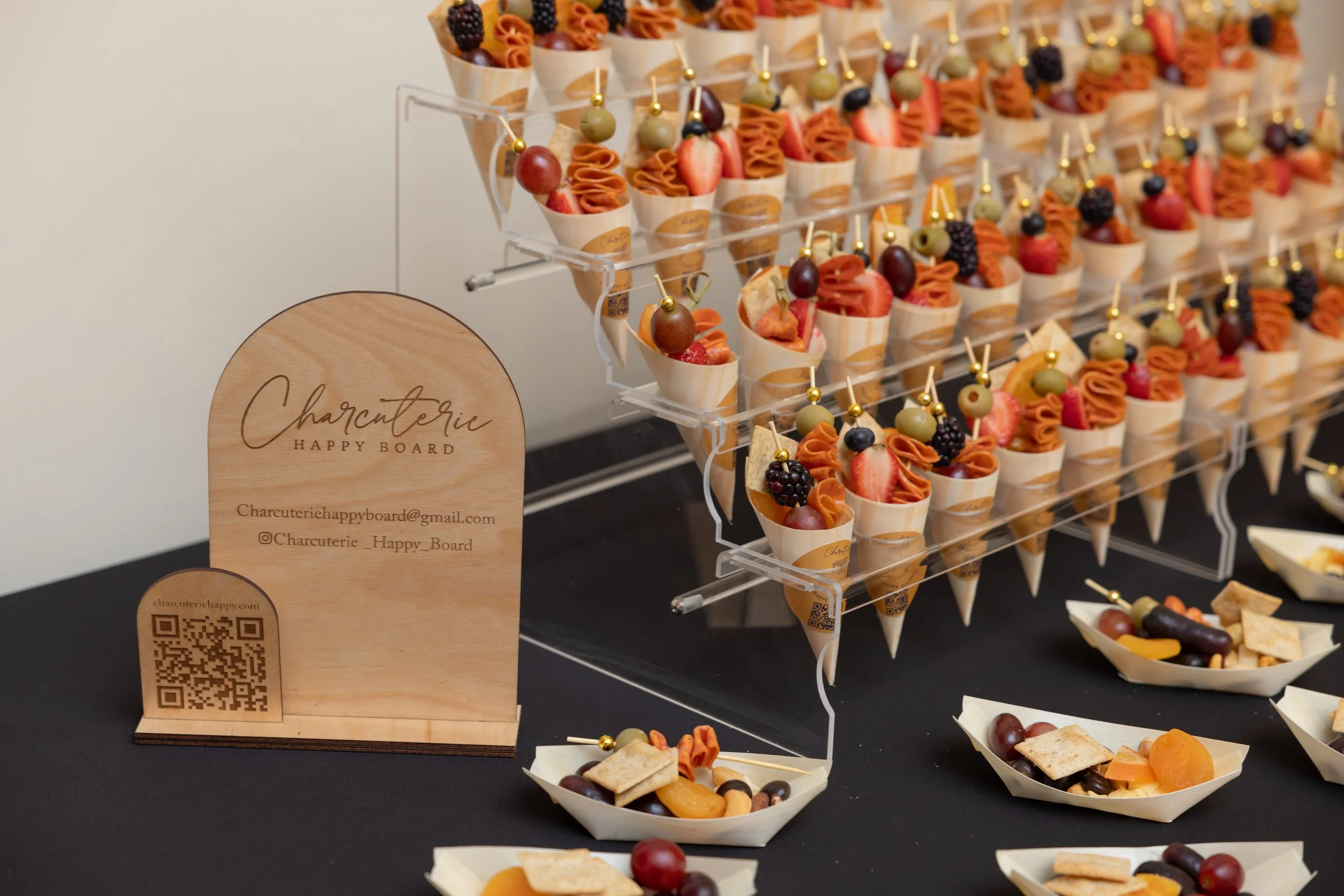 Assorted fruit and cheese appetizers on small cones and skewers displayed on a tiered acrylic stand at a party, with a wooden sign at the front that reads 'Charcuterie Happy Board' and includes contact information.