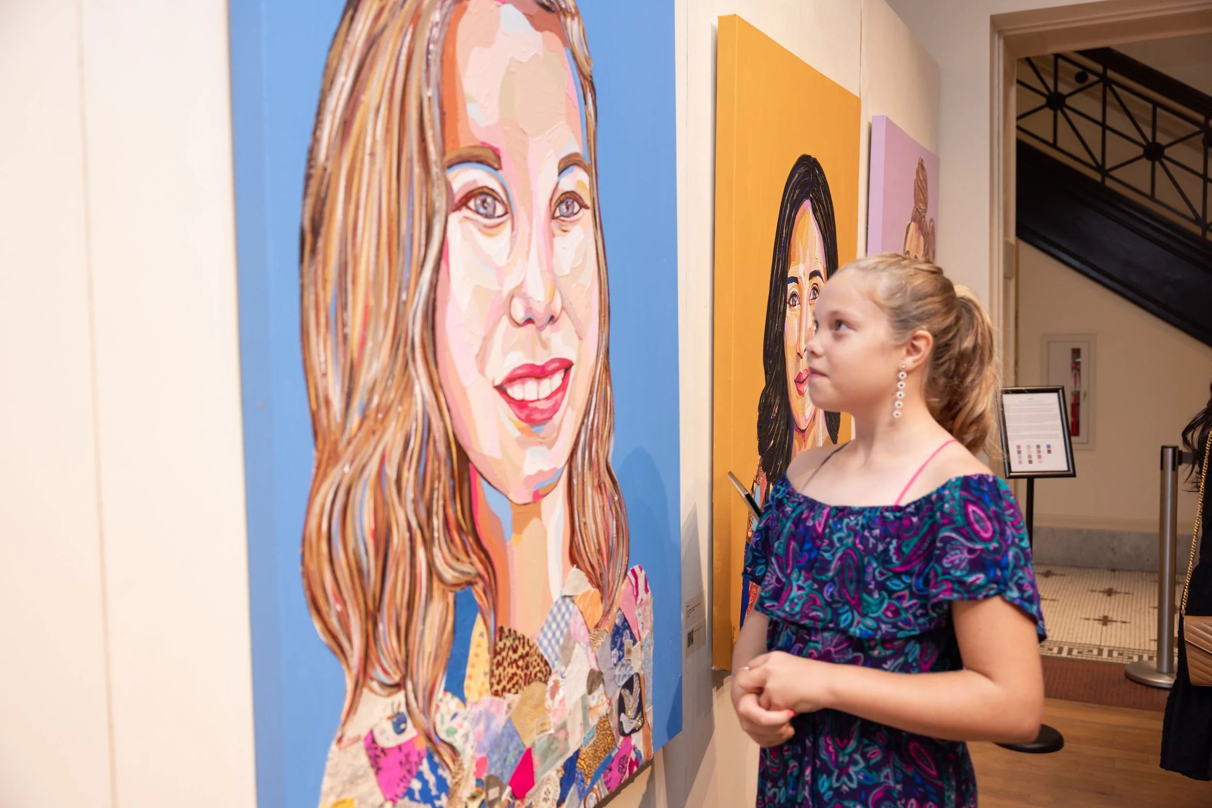 Young girl with a ponytail and earrings observing colorful portrait paintings in an art gallery.