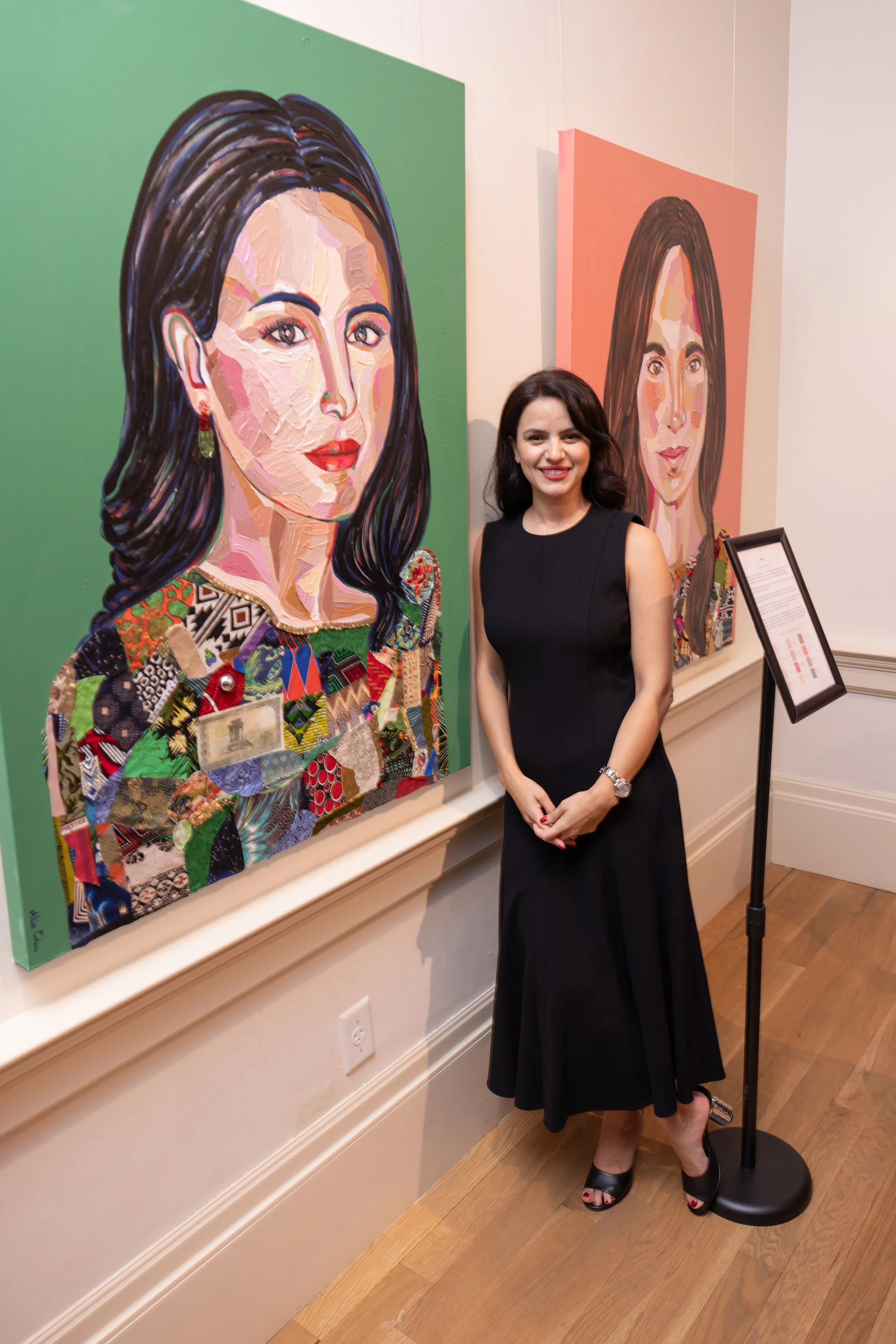 A woman in a black dress is standing next to a large portrait painting of a woman with dark hair and colorful makeup, displayed on an art gallery wall.