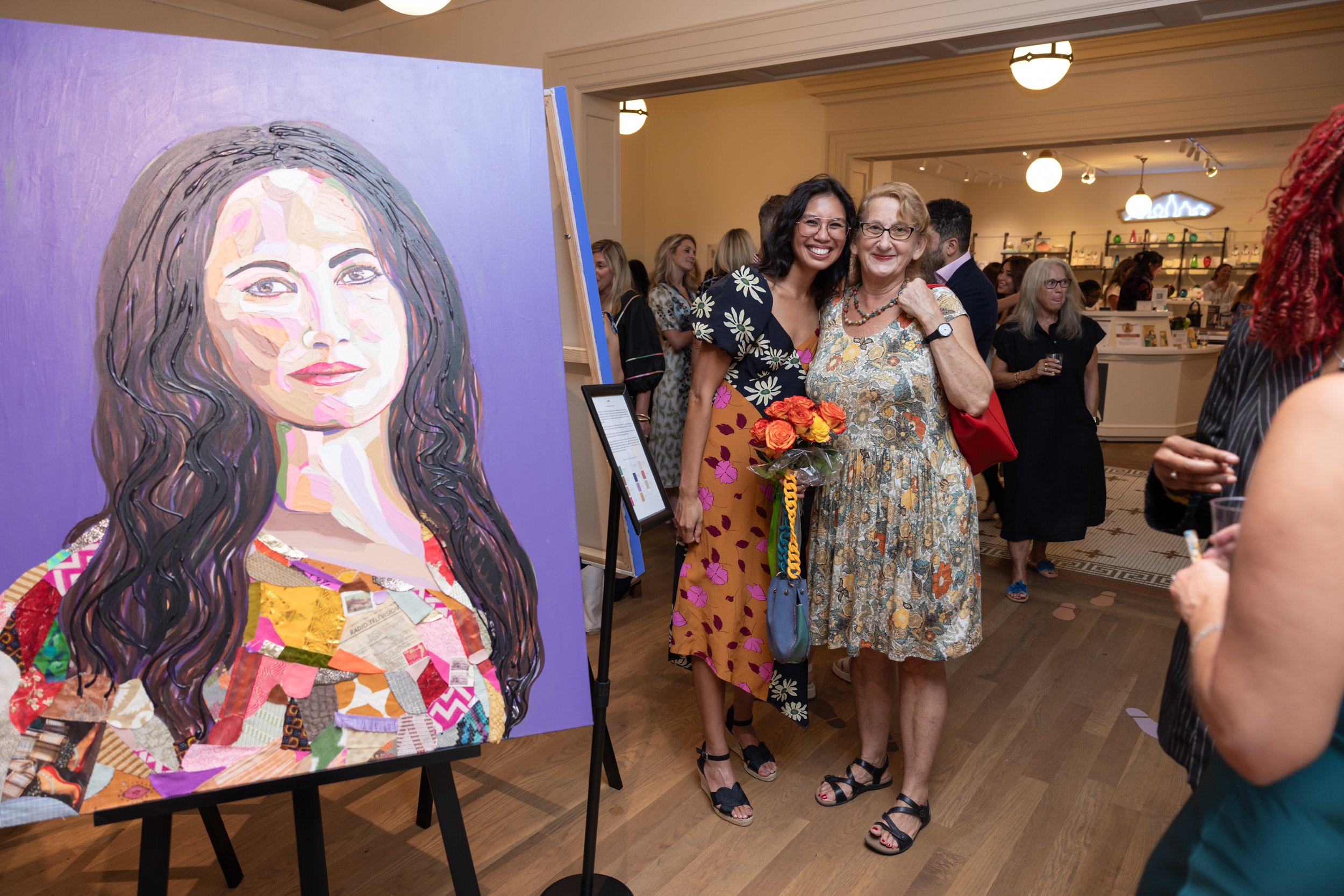 A colorful portrait of a woman with long dark hair displayed on an easel at an art gallery. People are mingling and viewing art in the background.