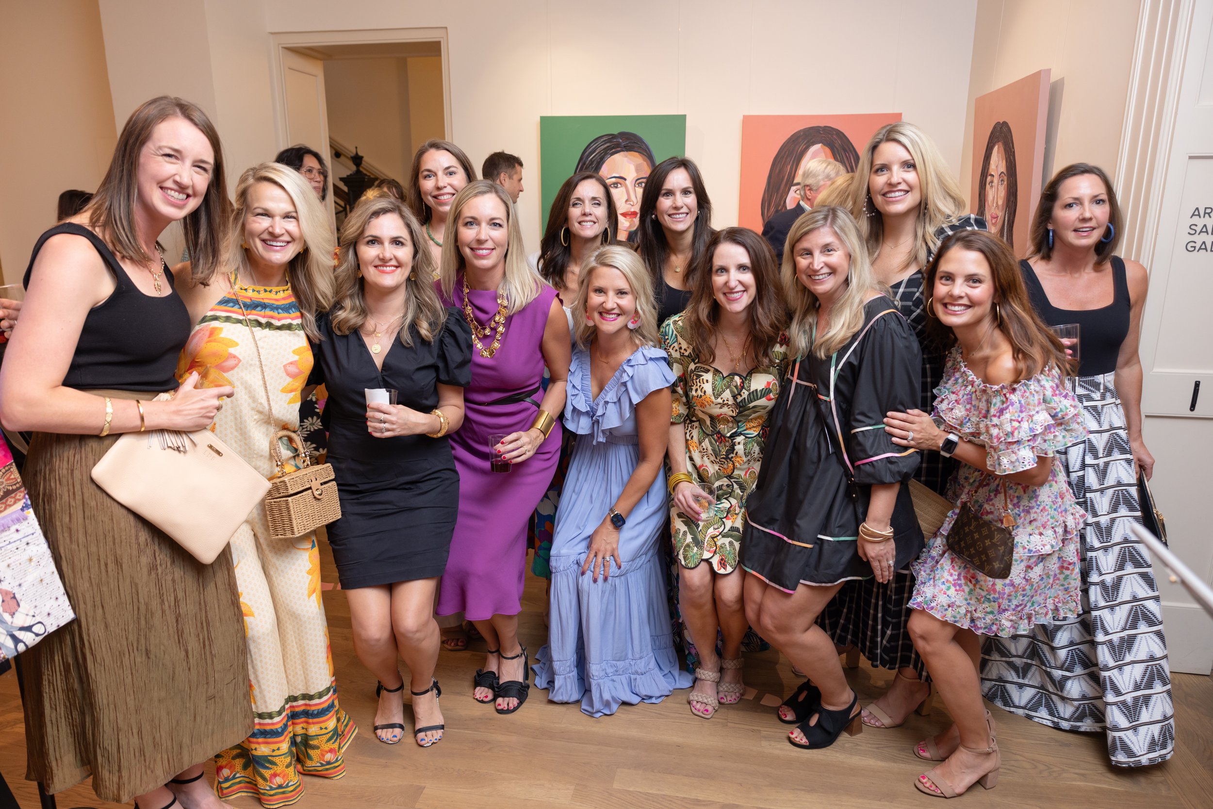 A group of women smiling and posing for a photo at an art gallery event. They are dressed in colorful and stylish outfits. The background shows paintings of women's faces on the wall.