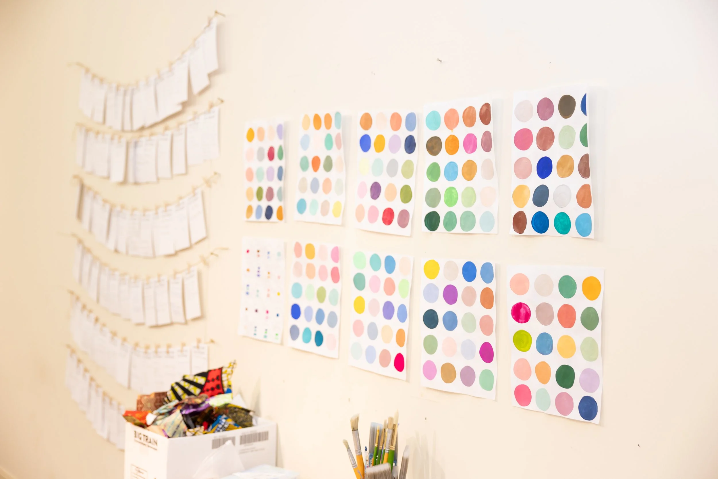 Color charts of colored circles arranged in rows and columns on a cream-colored wall, with paper flags hanging on strings and a box of candies below.