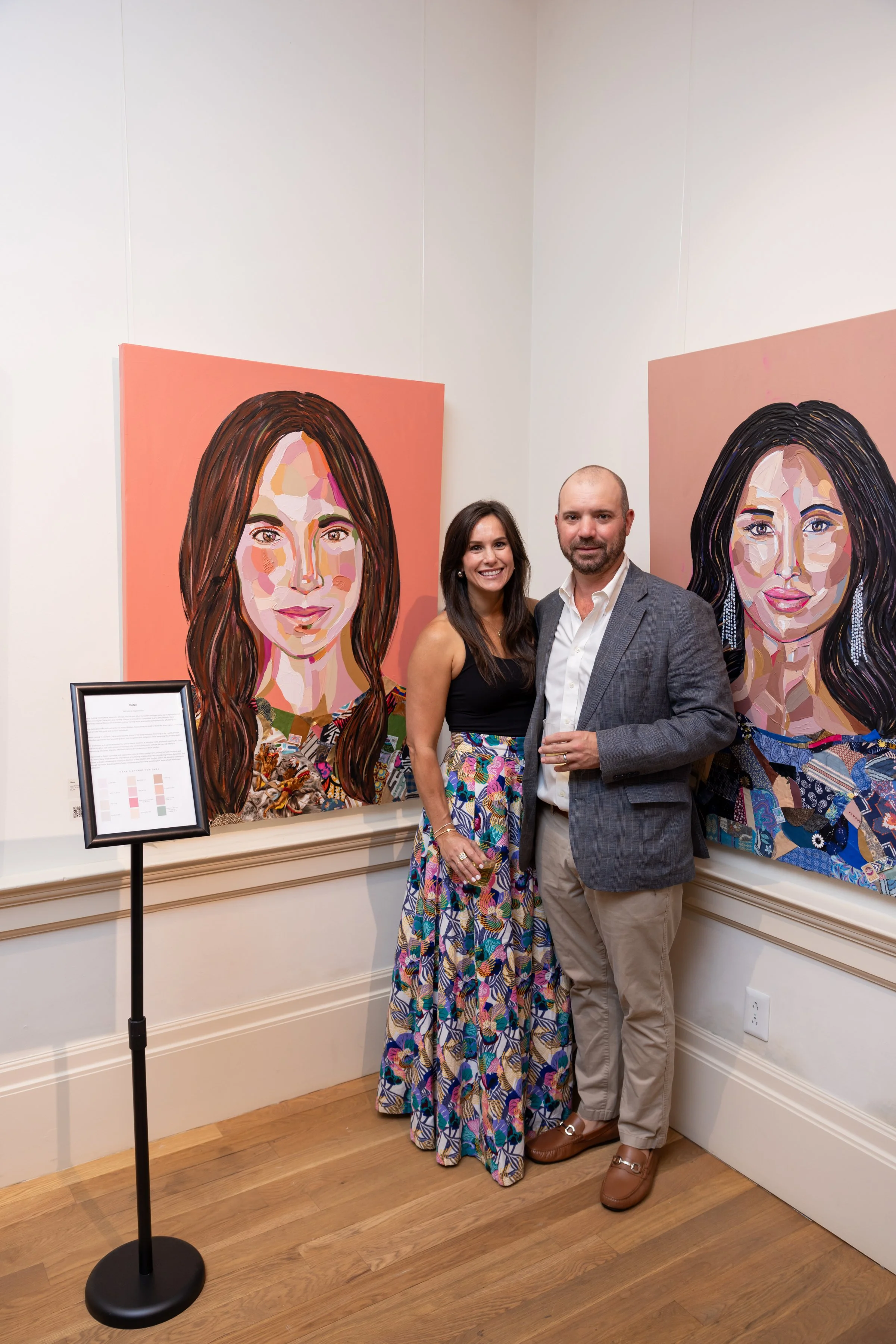 A man and a woman stand together in an art gallery, smiling at the camera. Behind them are two large colorful paintings of women with different hairstyles. The woman is wearing a black top and a floral skirt, and the man is in a gray blazer, white sh