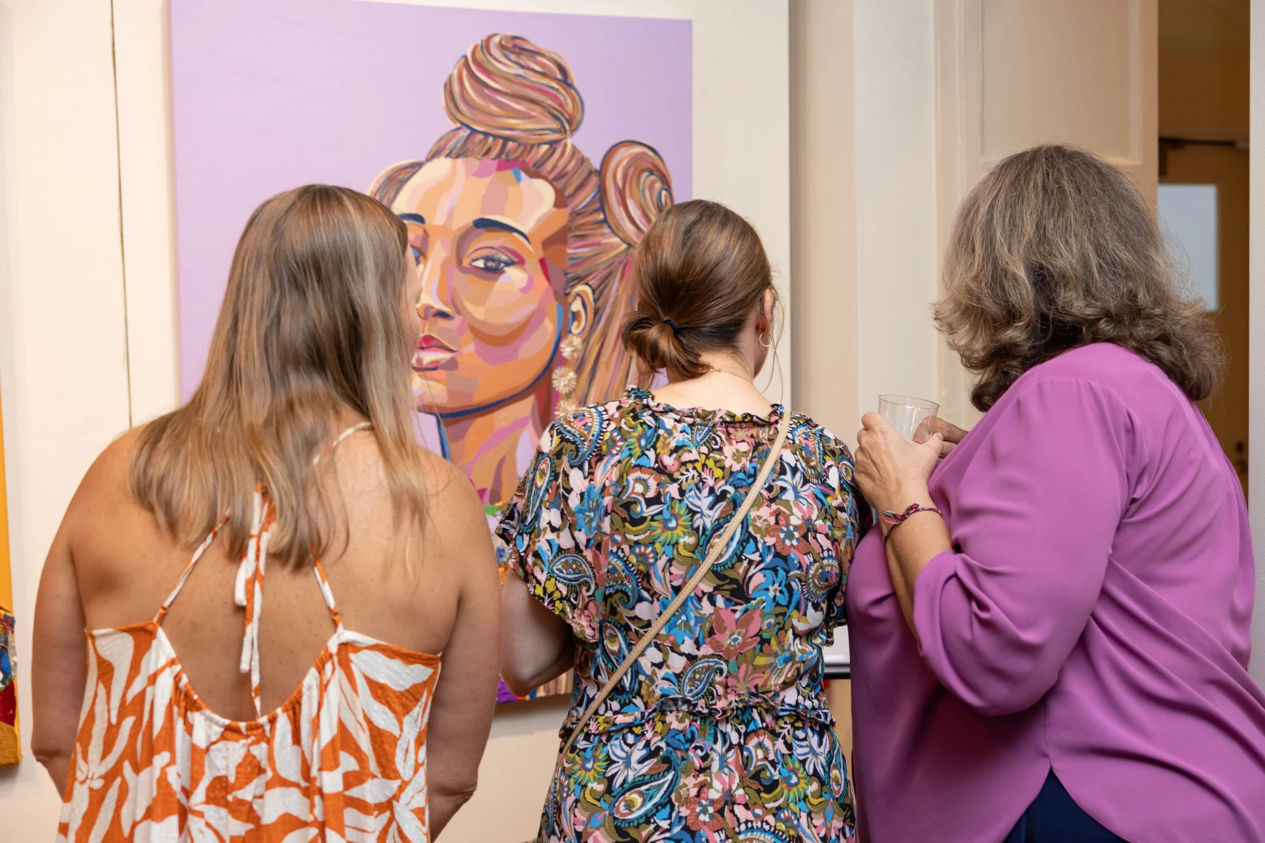 Three women with varied hair colors and styles viewing a large colorful portrait of a woman with styled hair wearing earrings.