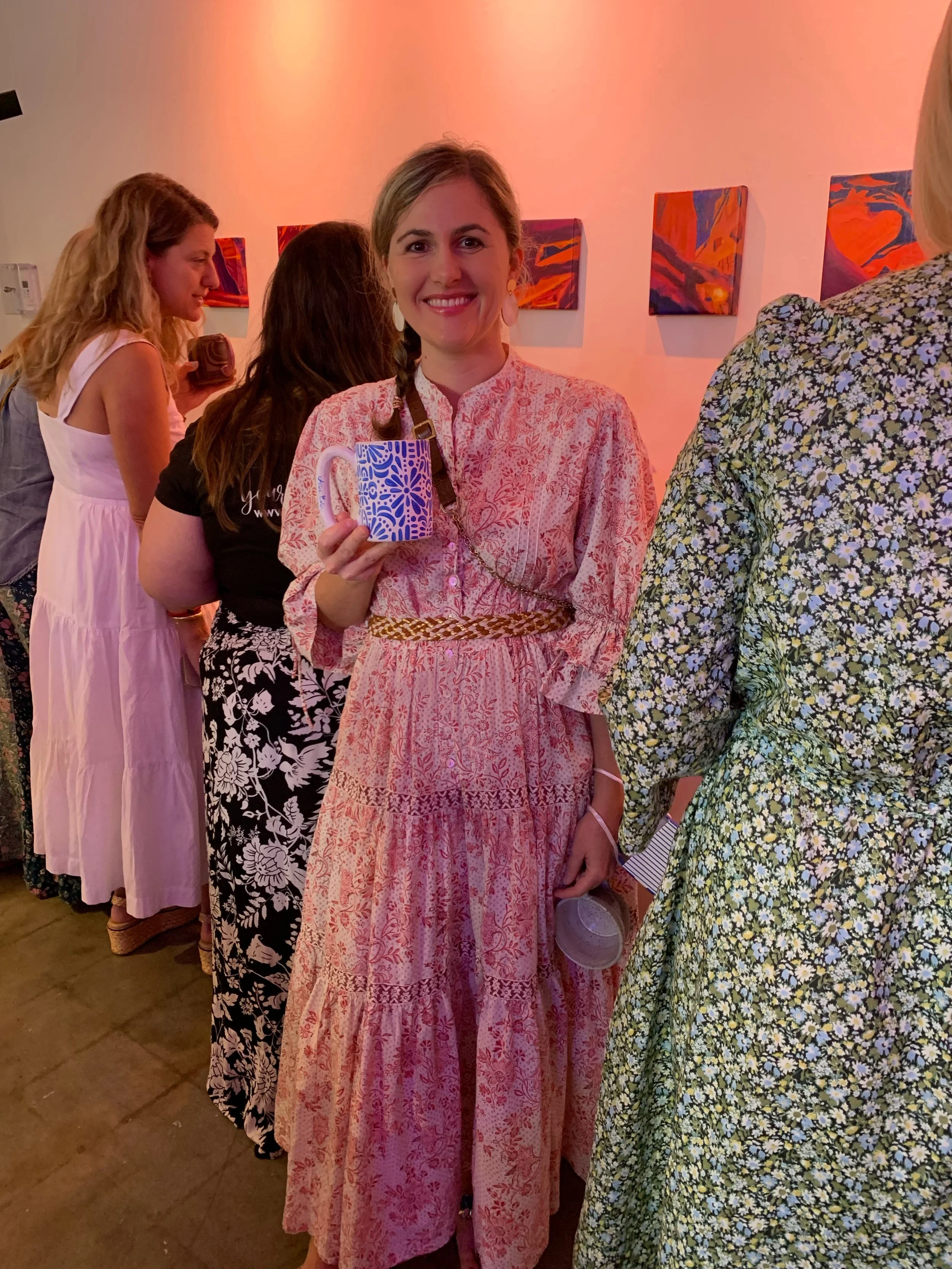 A smiling woman holding a blue and white patterned mug, standing in an art gallery with colorful abstract artworks on the wall, surrounded by other people in floral and pastel dresses.