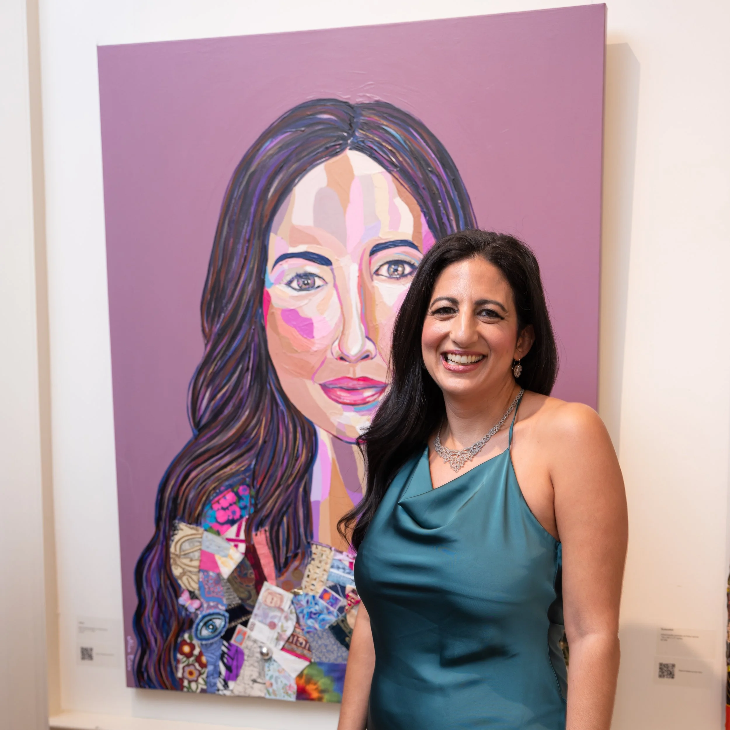 Woman with dark hair smiling in front of a colorful portrait painting of a woman's face displayed on a wall.
