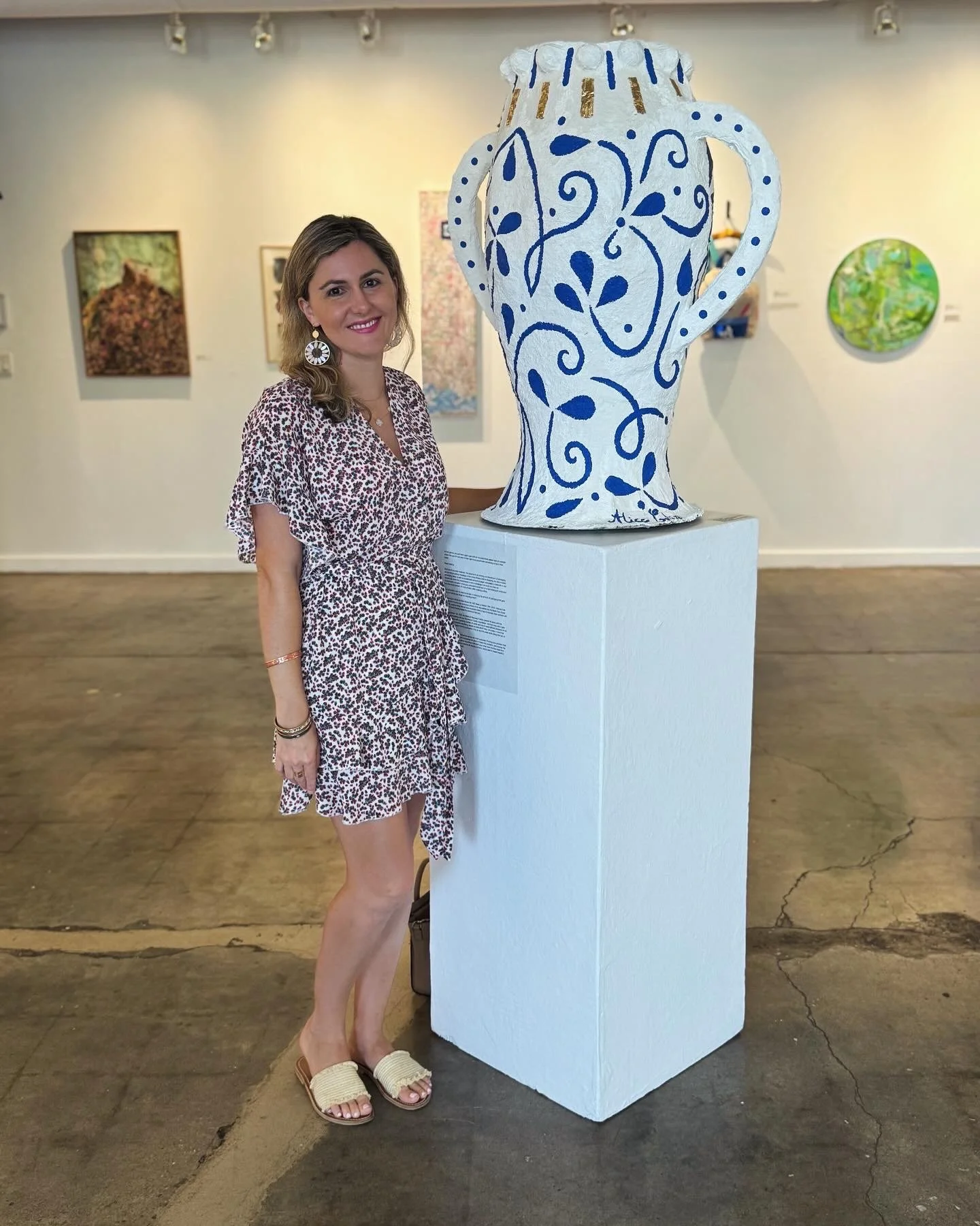 A woman with light brown hair wearing a leopard print dress and earrings, standing next to a large white and blue ceramic-style vase sculpture on display in an art gallery.
