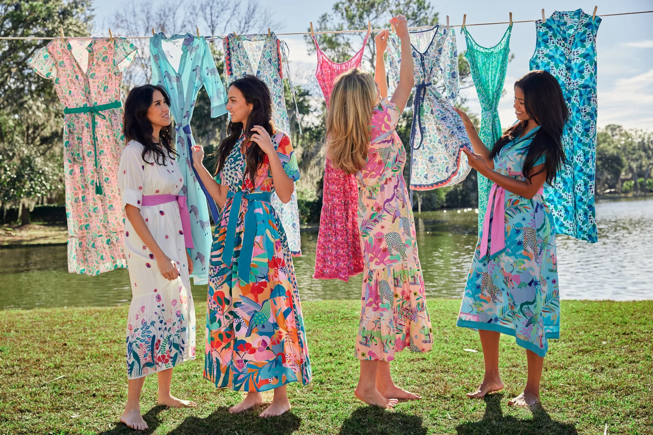 Five women standing barefoot outdoors in a park, hanging colorful dresses on a clothesline, with trees and a body of water in the background.