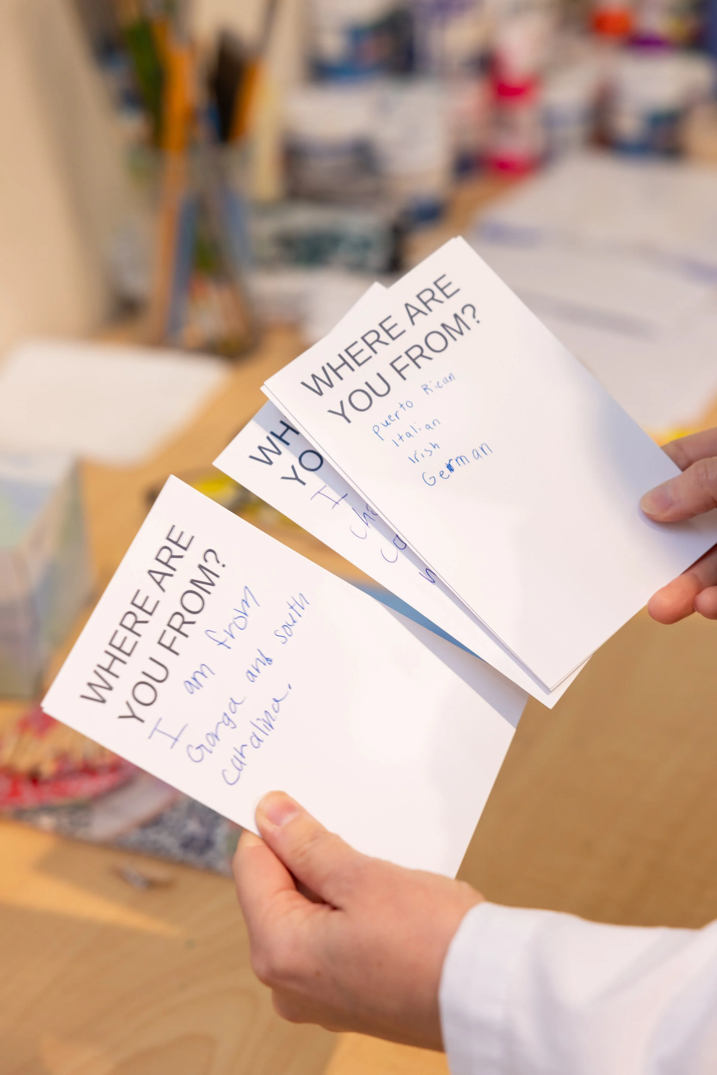 A person holding three sheets of paper with the question 'Where are you from?' printed at the top. The sheets have handwritten responses indicating places such as Puerto Rican, Italian, Irish, and German. In the background, there is a blurred display of art supplies.