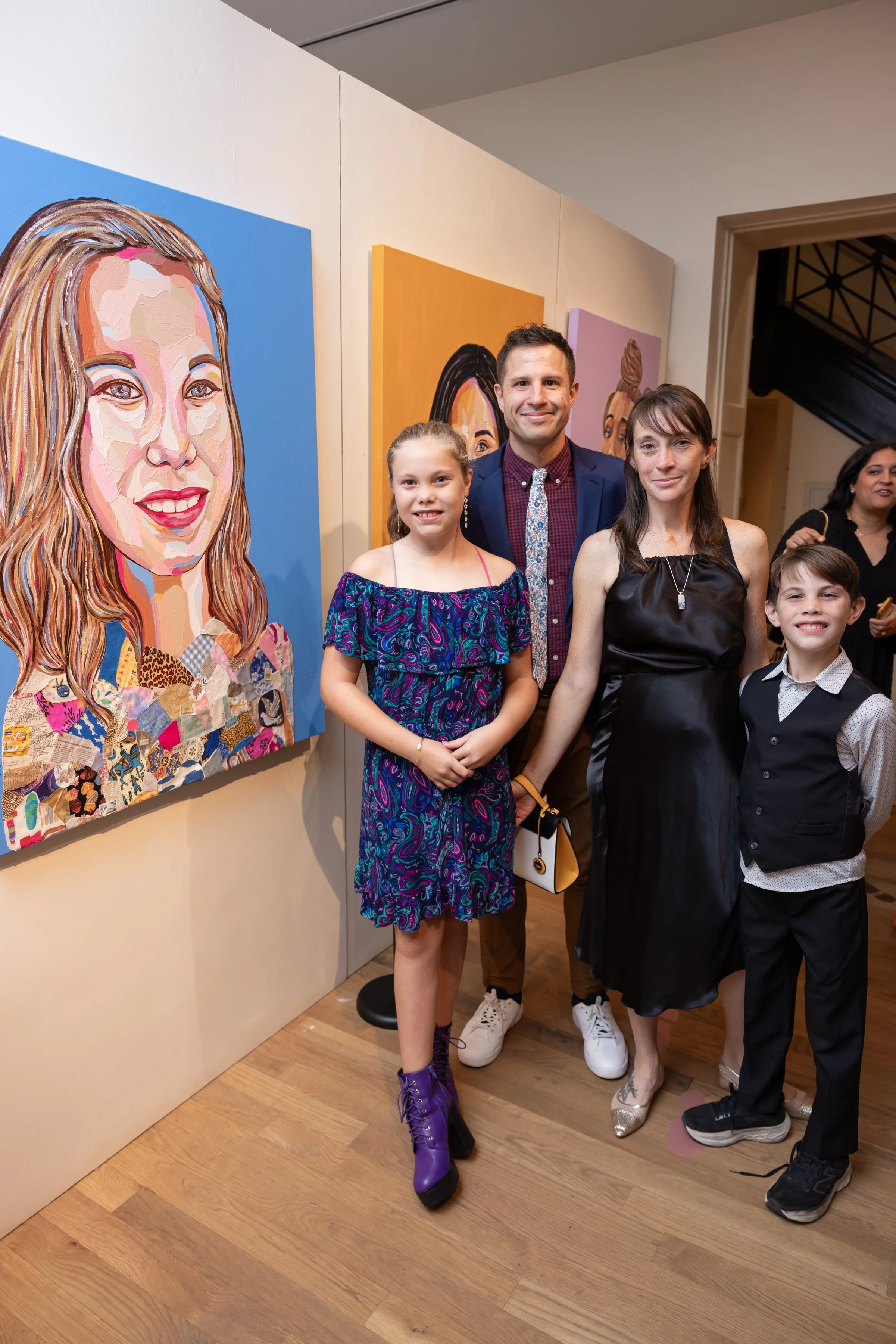 A group of five people, three women and two children, standing in front of colorful portrait paintings at an art gallery. They are dressed in stylish clothing and smiling at the camera.