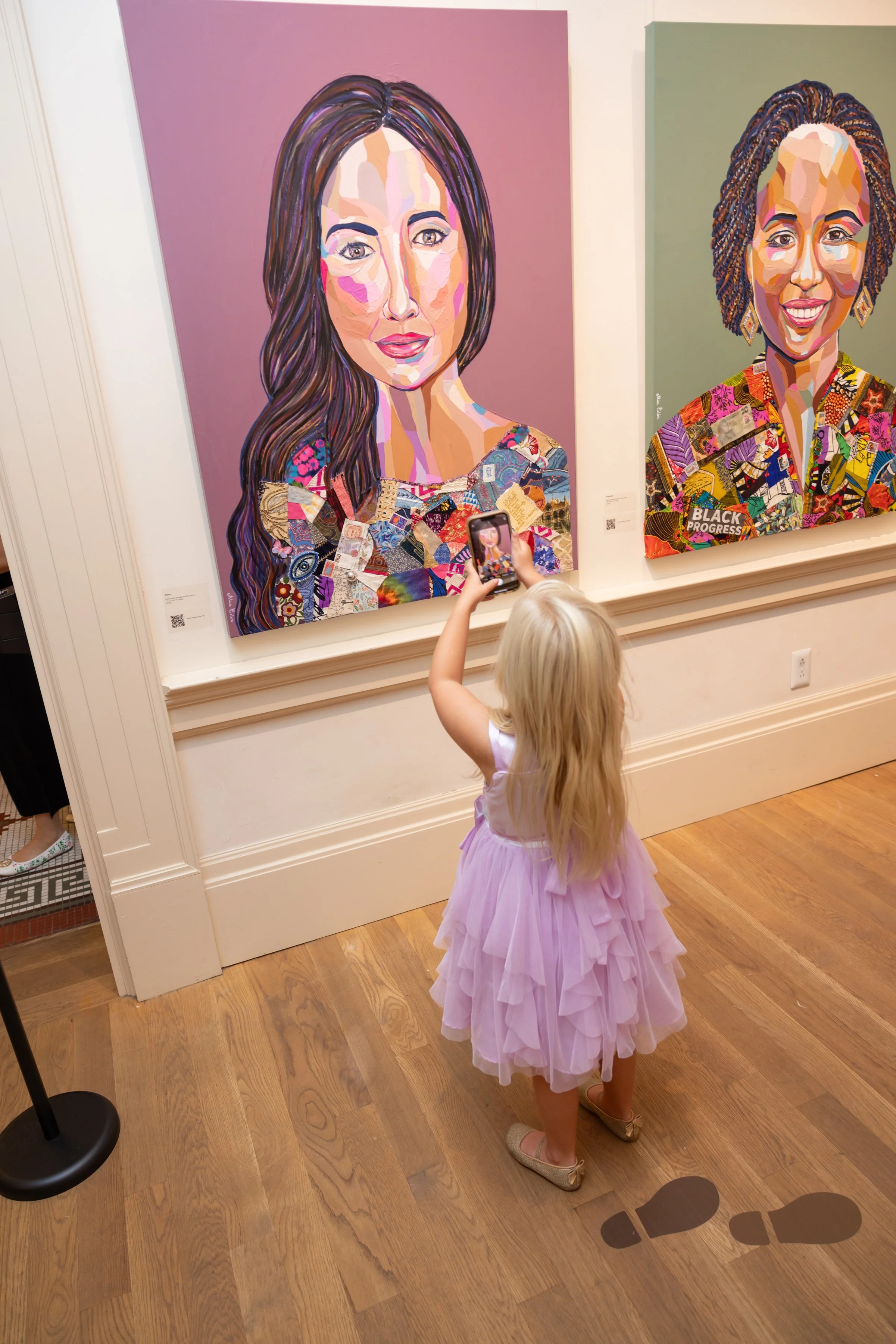 A young girl with long blonde hair wearing a purple ruffled dress is taking a photo with her smartphone of two large colorful portraits displayed on gallery walls. The portraits feature women with vibrant, collage-style artistic renditions of their f
