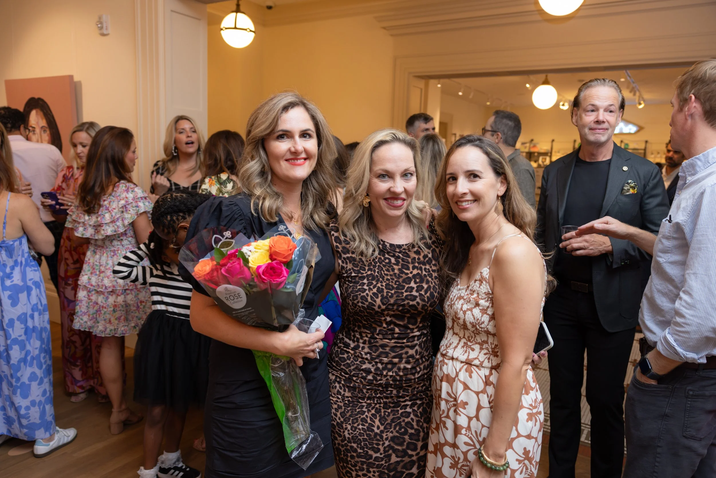People gathered at a social event with three women in the foreground, one holding a bouquet of flowers. The background has a group of people conversing and interacting in a warmly lit room.