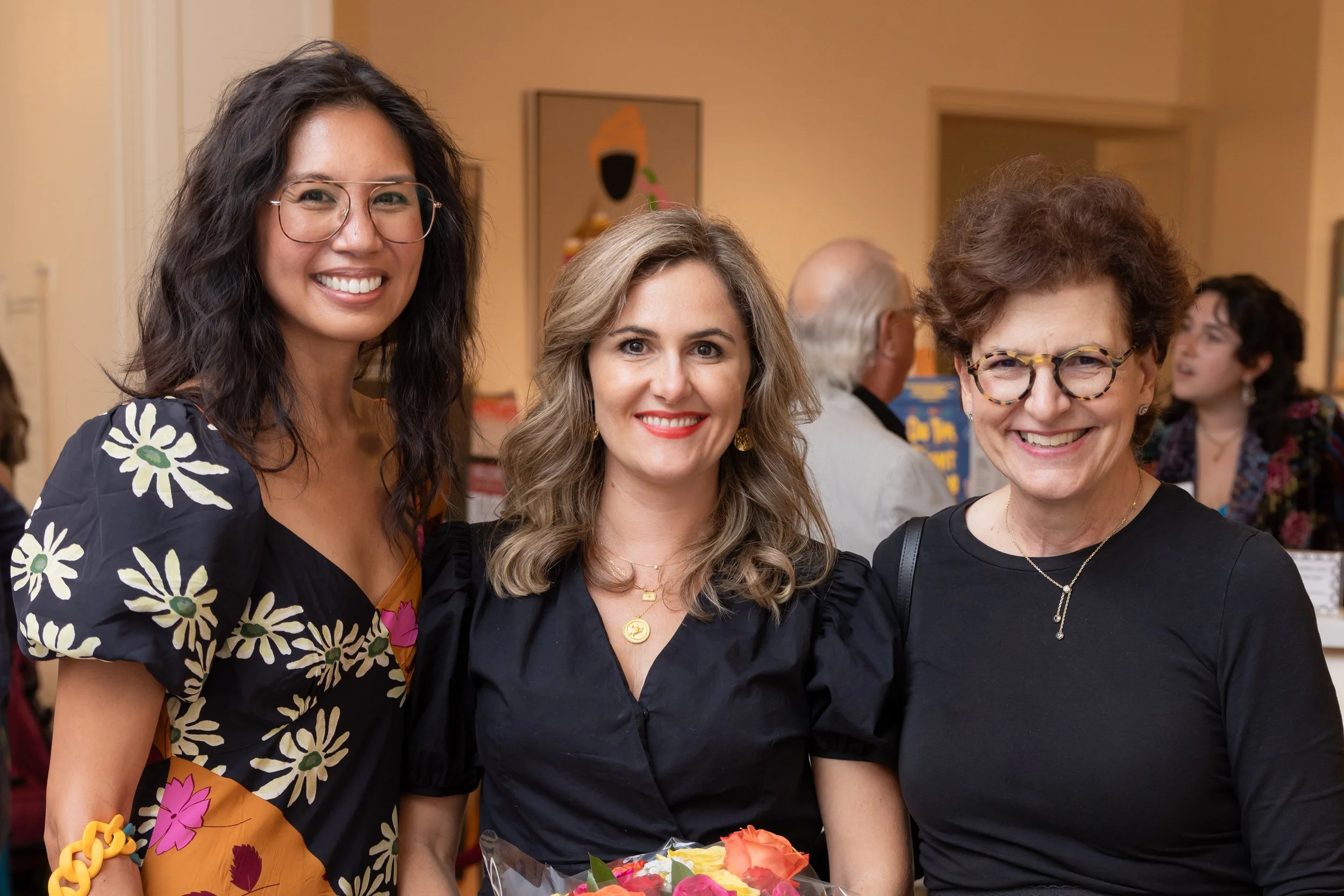 Three women smiling at a social event, with a table of flowers in front of them, in a warmly lit room.