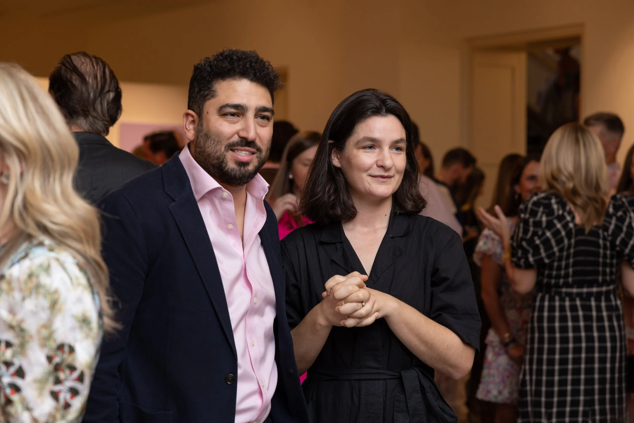 A man and a woman are holding hands and engaging in conversation at a social event with many attendees in the background.
