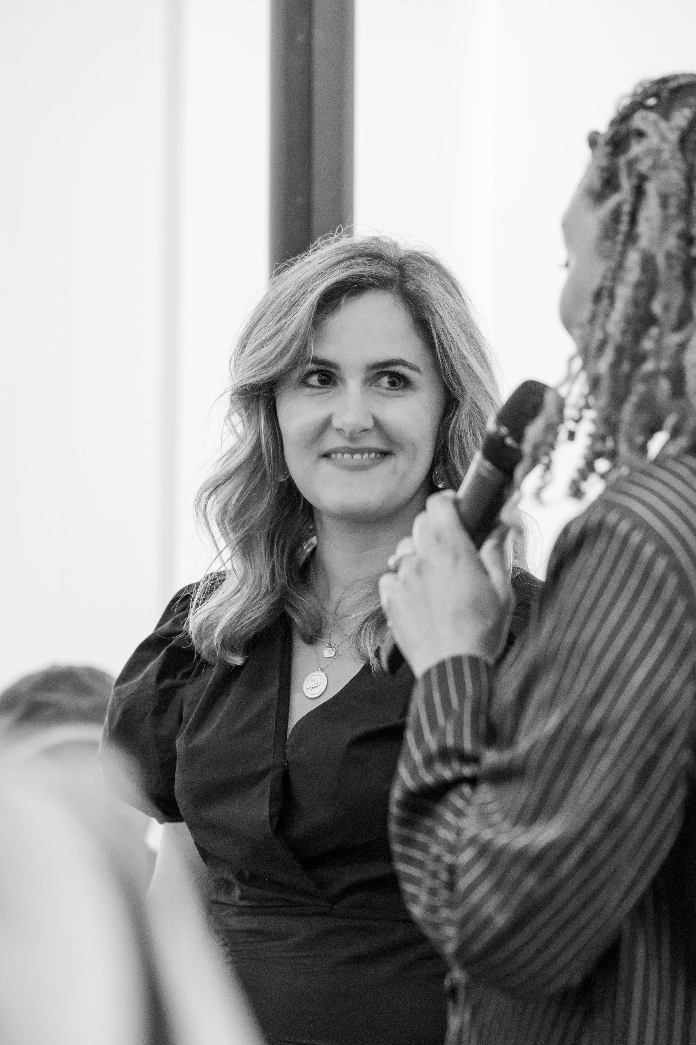 Two women talking, one holding a microphone. The woman on the left is smiling with wavy hair, wearing a dark blouse and jewelry. The woman on the right is partially visible, holding the microphone, wearing a striped shirt. The setting appears to be a