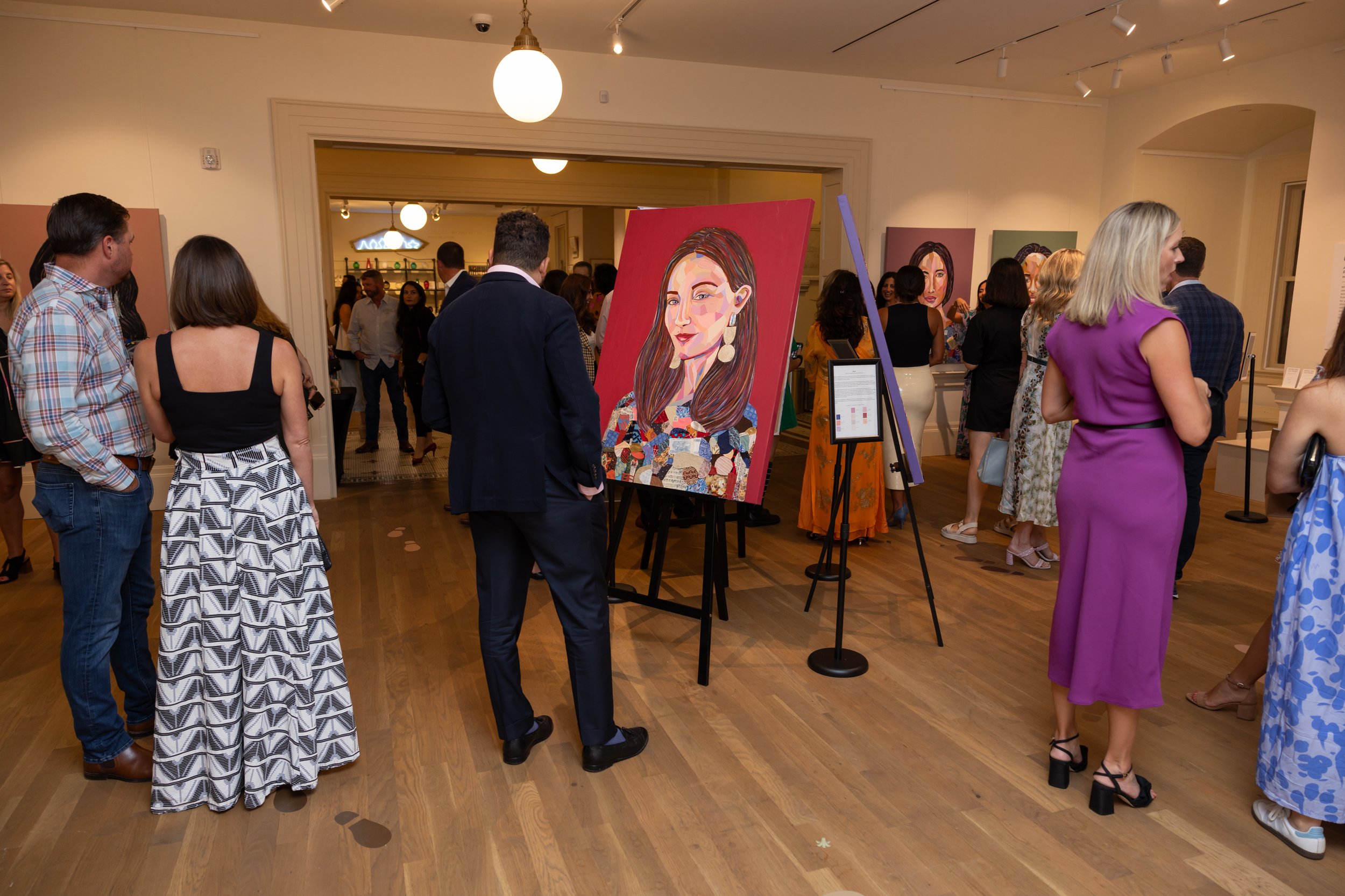 Gallery event with people viewing colorful portraits of women on display, some engaging in conversations inside a well-lit art gallery with wooden floors.