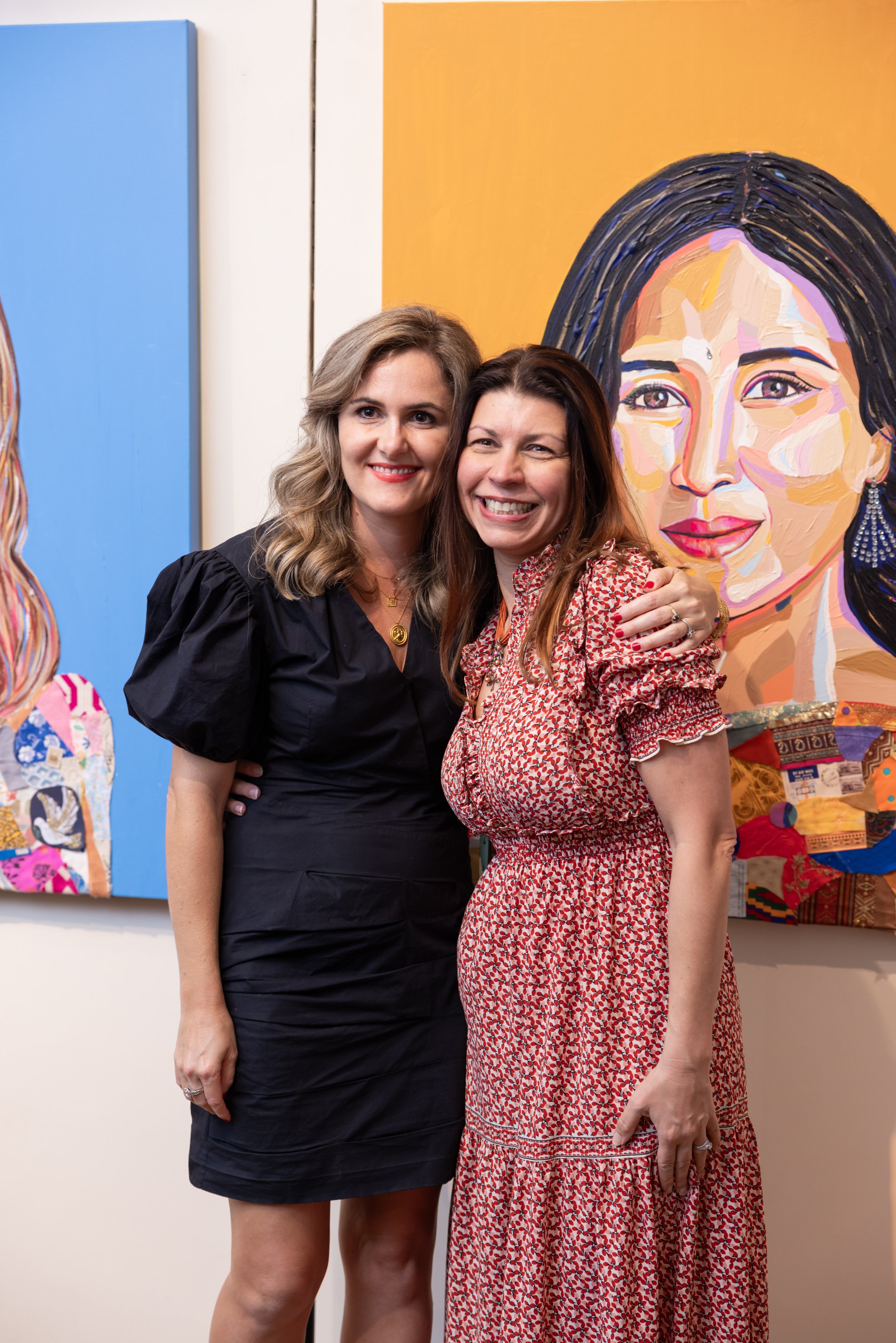 Two women standing together in front of colorful abstract portraits at an art gallery, smiling and hugging.