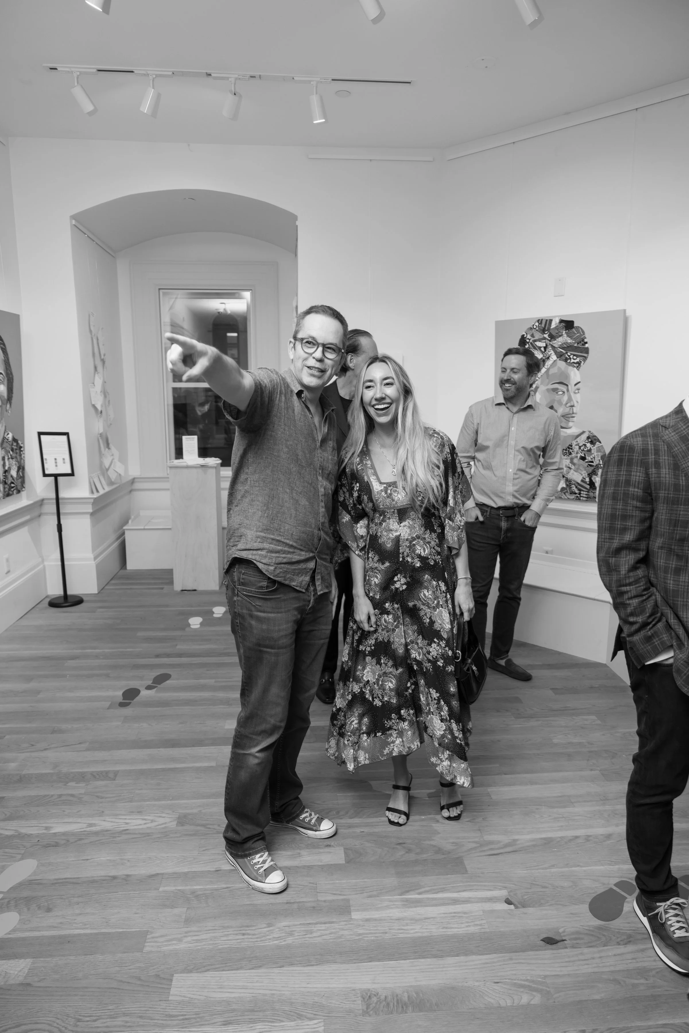 Group of five people in an art gallery, two women and three men, smiling and talking. One man points to something outside the frame. The background includes colorful art on white walls.