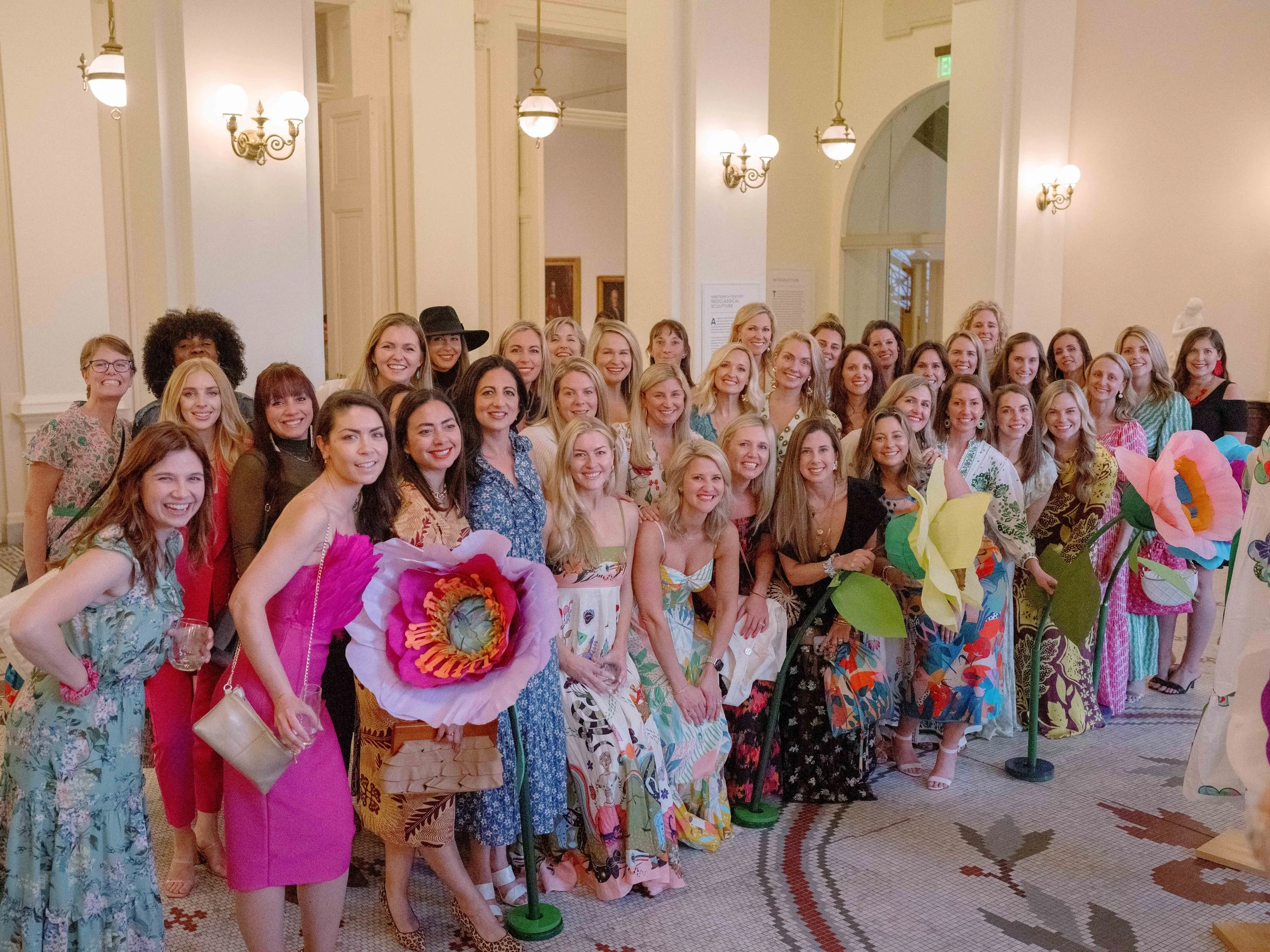 Group of women dressed in colorful, floral dresses posing for a photo inside a historic building with ornate lighting and tiled floors.