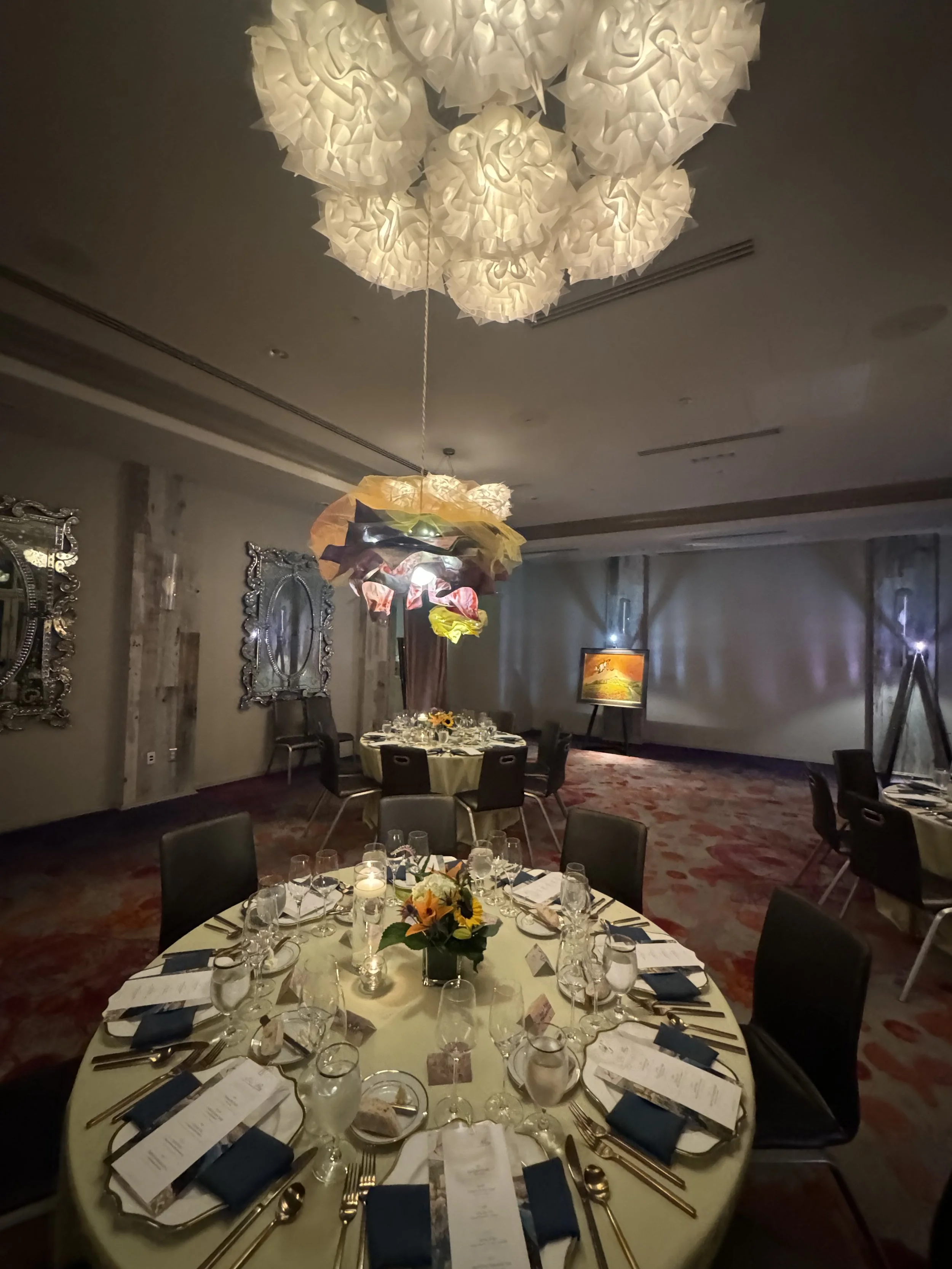 Elegant banquet hall with round tables set for a formal dinner, featuring candles, floral arrangements, and neatly arranged dinnerware. Modern hanging light fixtures decorate the ceiling.