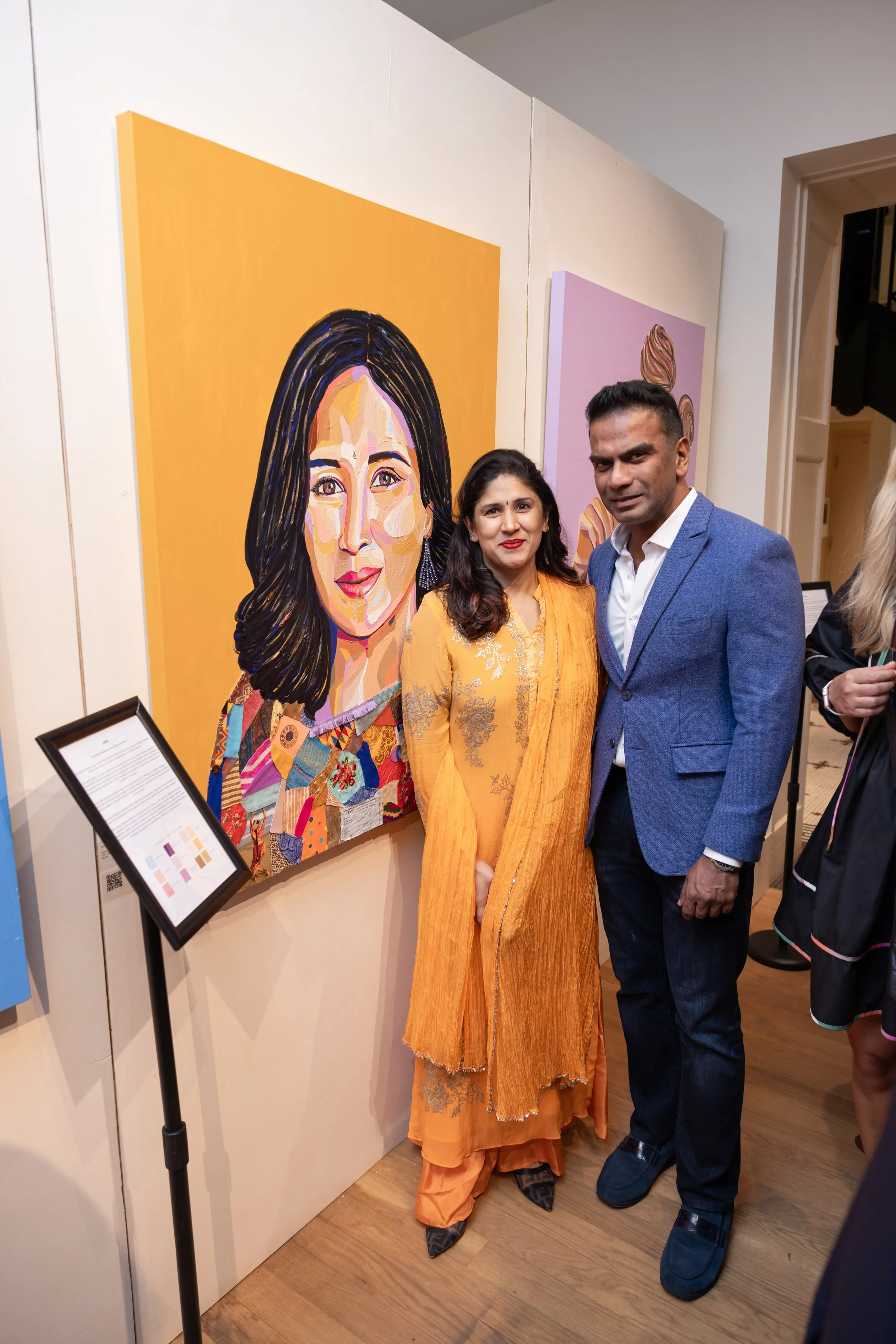 Two people posing in front of colorful pop art portraits at an art gallery. The woman is wearing a yellow dress with embroidery, and the man is in a blue suit and white shirt.