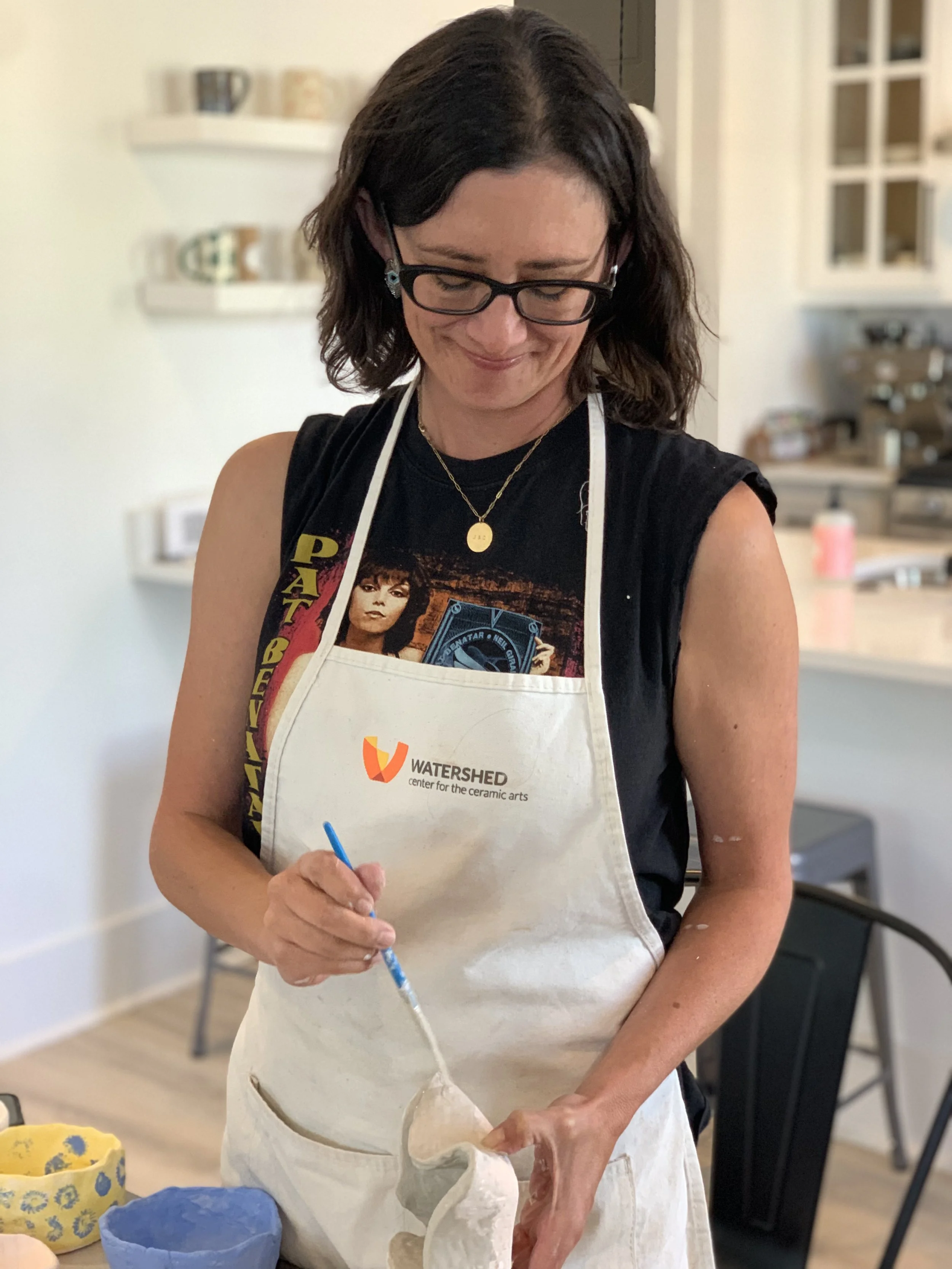 A woman with shoulder-length brown hair, wearing glasses, a black T-shirt with a graphic of a woman, and a white apron, is working on a ceramic project in a well-lit kitchen or studio. She is holding a blue sculpting tool and is shaping a white ceramic piece. In the background, there are shelves with mugs and containers, and a counter with various equipment.
